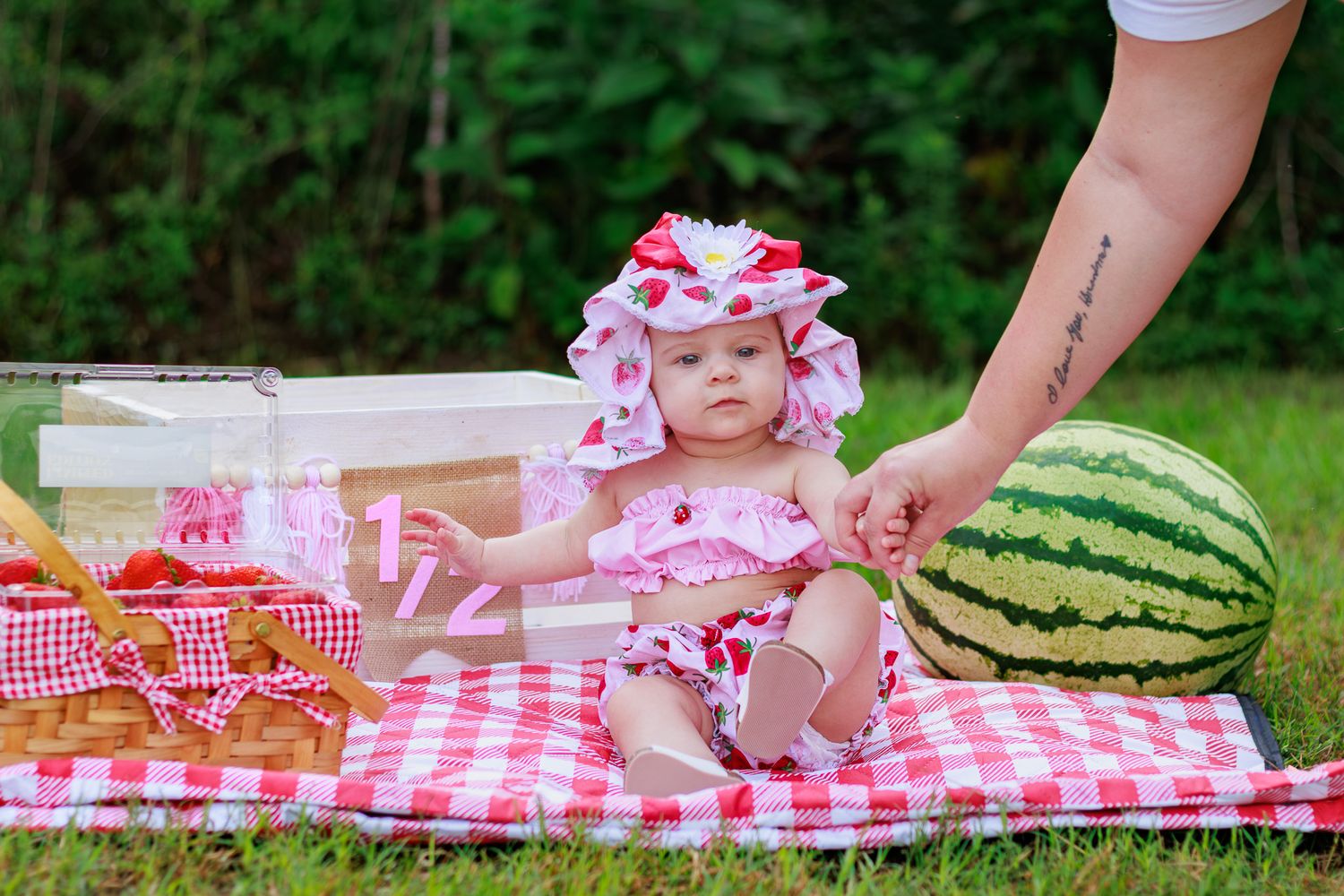 Adorable baby in red gingham set holds adult hand on picnic blanket beside large watermelon outdoors.