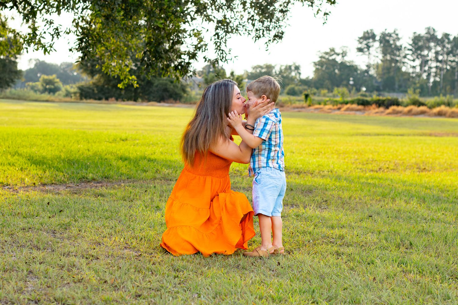 A mother in an orange dress kneels on green grass sharing a tender moment with her young son on a sunny day.