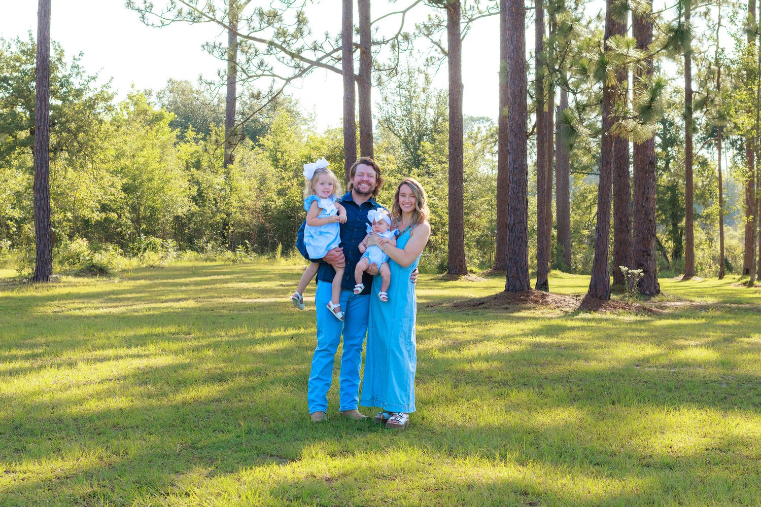 Happy family with two young children posing outdoors in a sunny green park surrounded by tall pine trees.