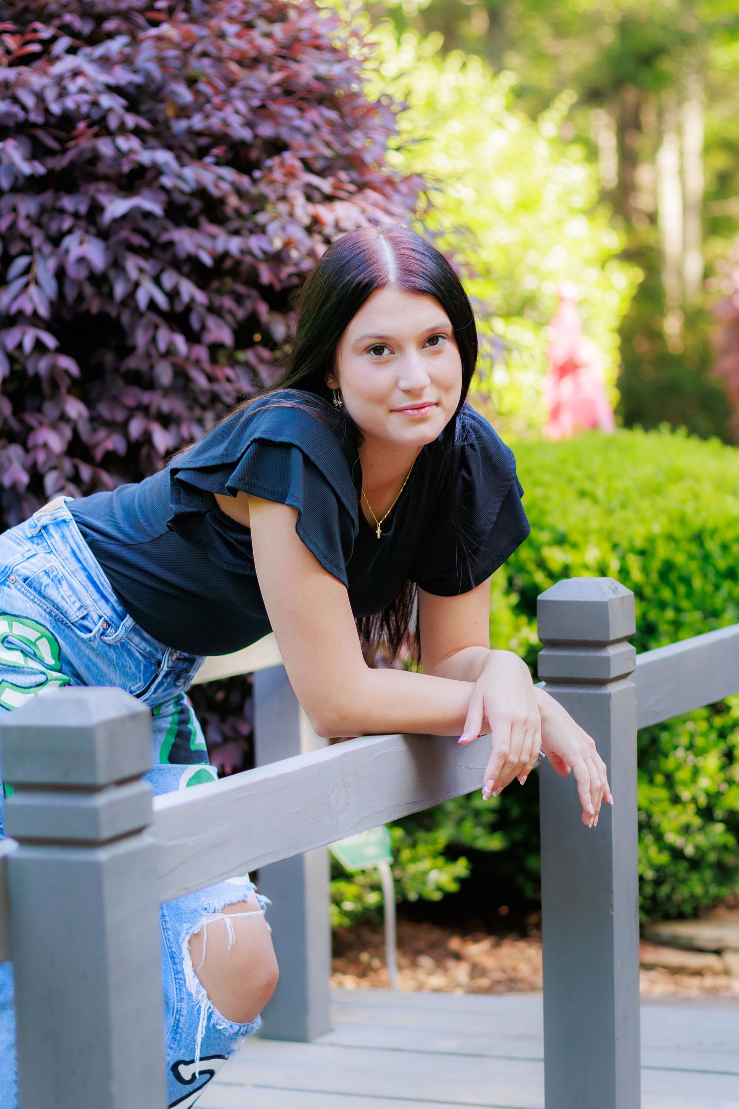 Young woman in black top and ripped jeans leaning on gray deck railing in lush green garden.