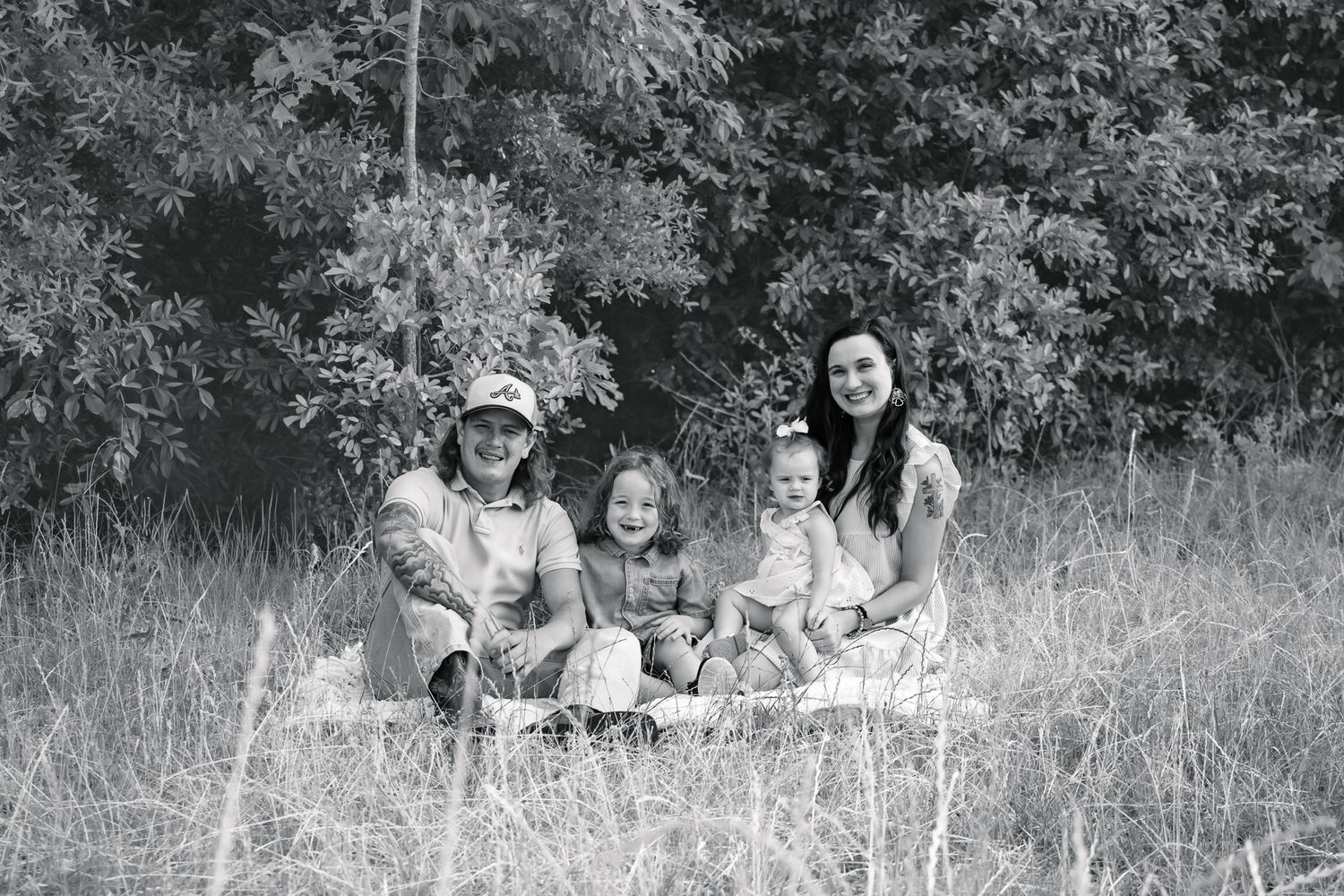 Black and white family portrait of four people sitting in a meadow with tall grass and trees in the background.
