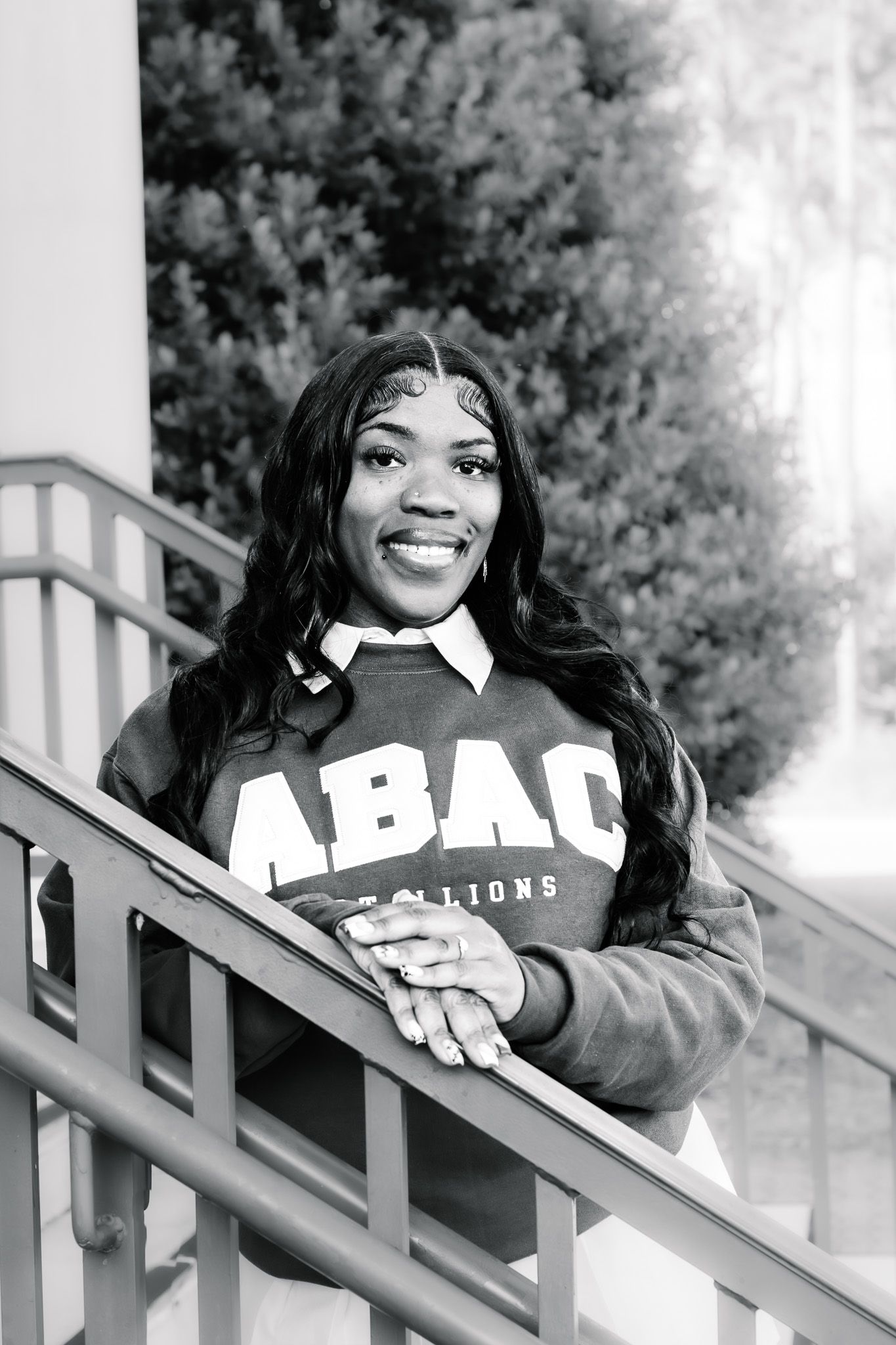 Smiling young woman in ABAC Stallions sweatshirt leaning on stair railing outdoors in black and white photo.