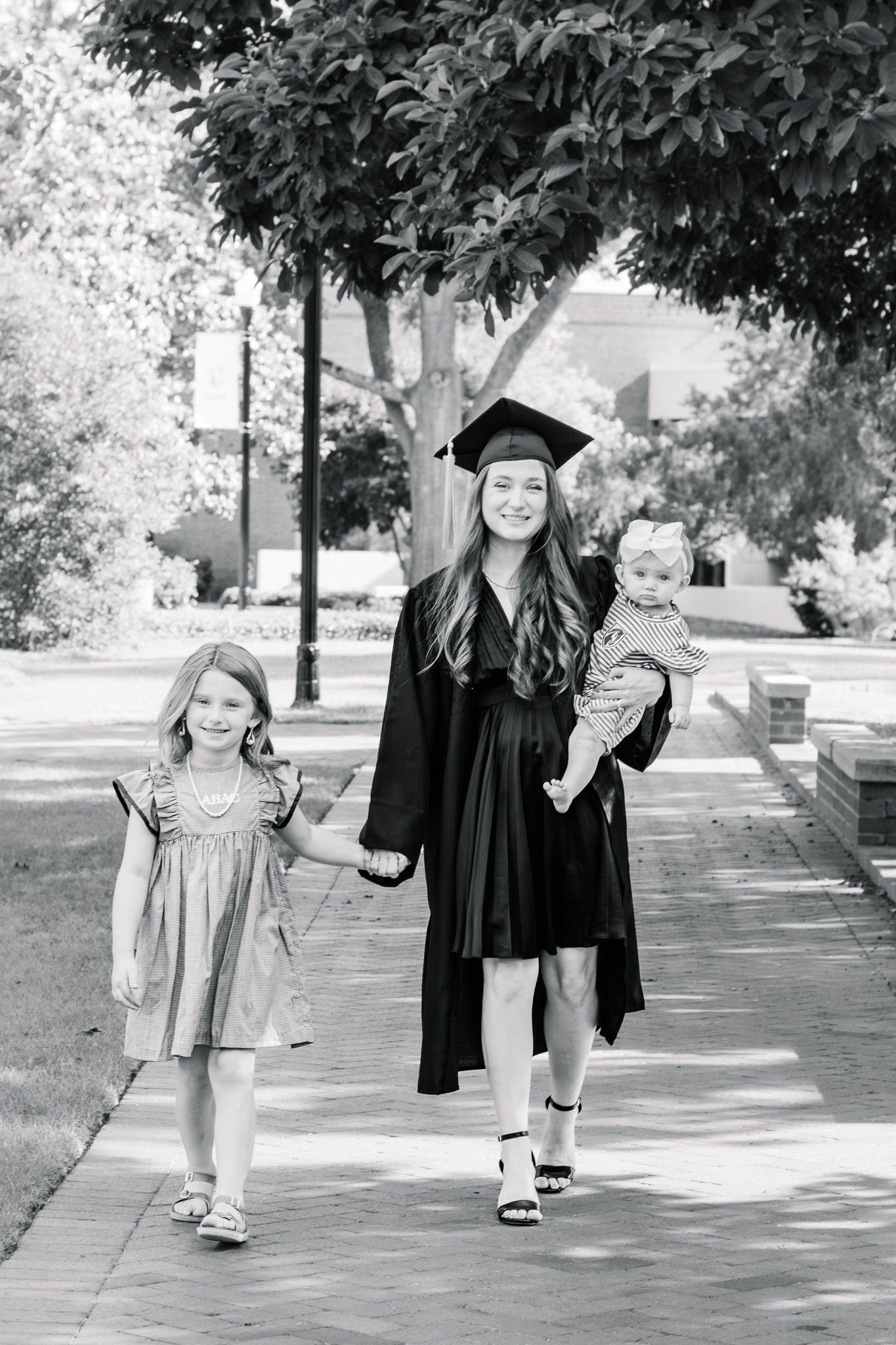 Graduate in cap and gown walks with two young children on a sunny campus pathway in black and white photo.
