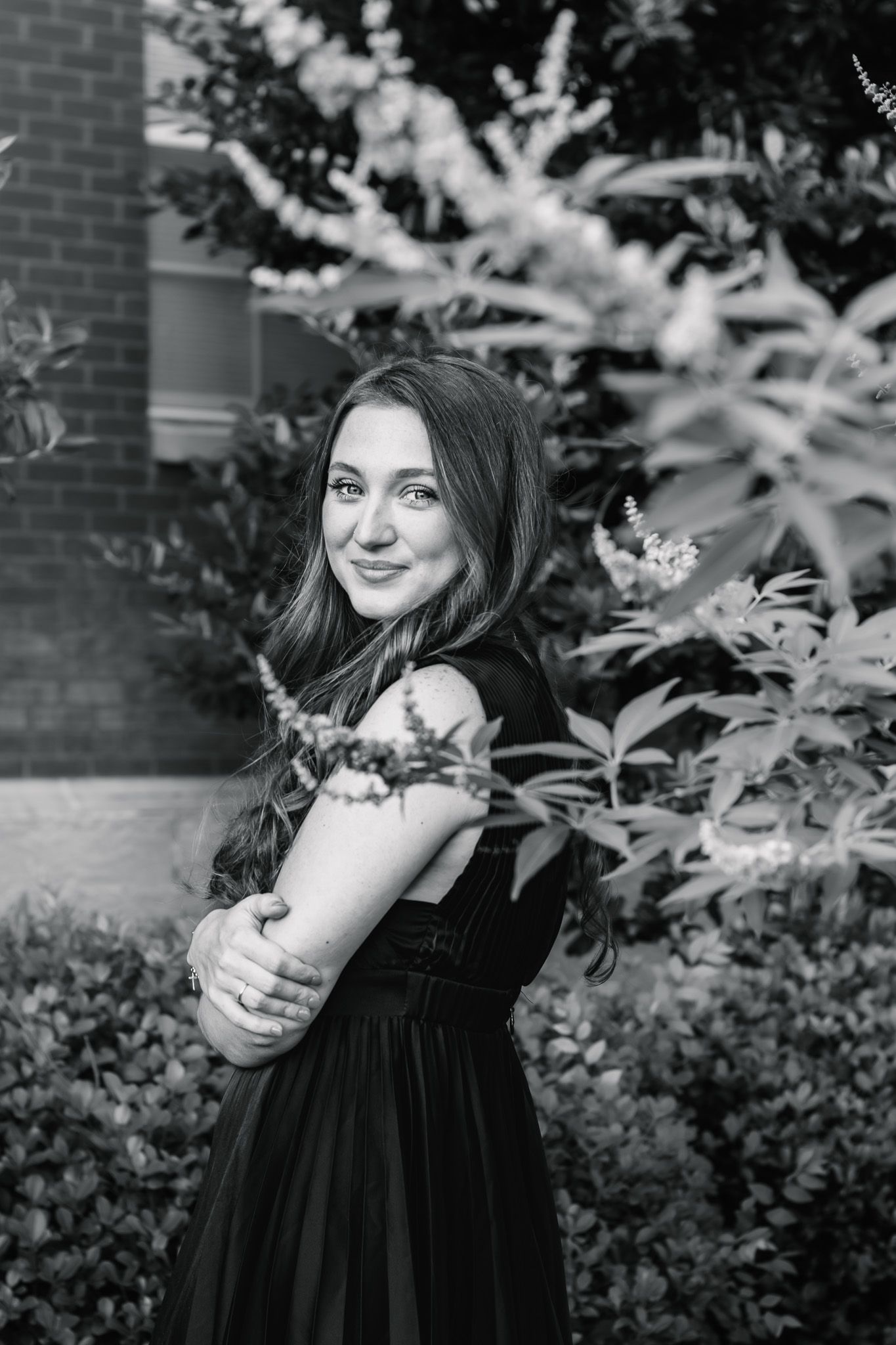 Young woman in black dress poses gracefully among garden shrubs in black and white portrait.