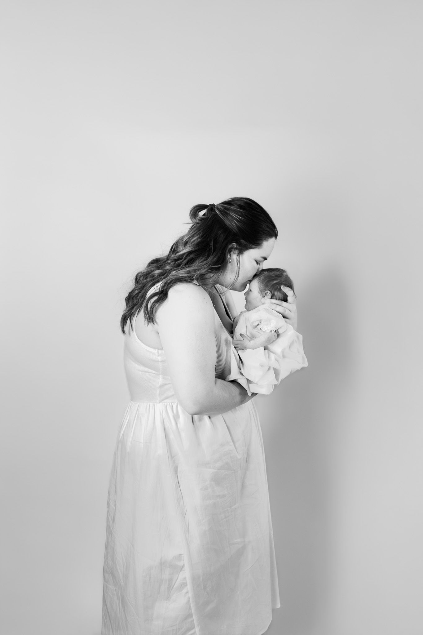 Black and white photo of a mother in white dress tenderly kissing her newborn baby.