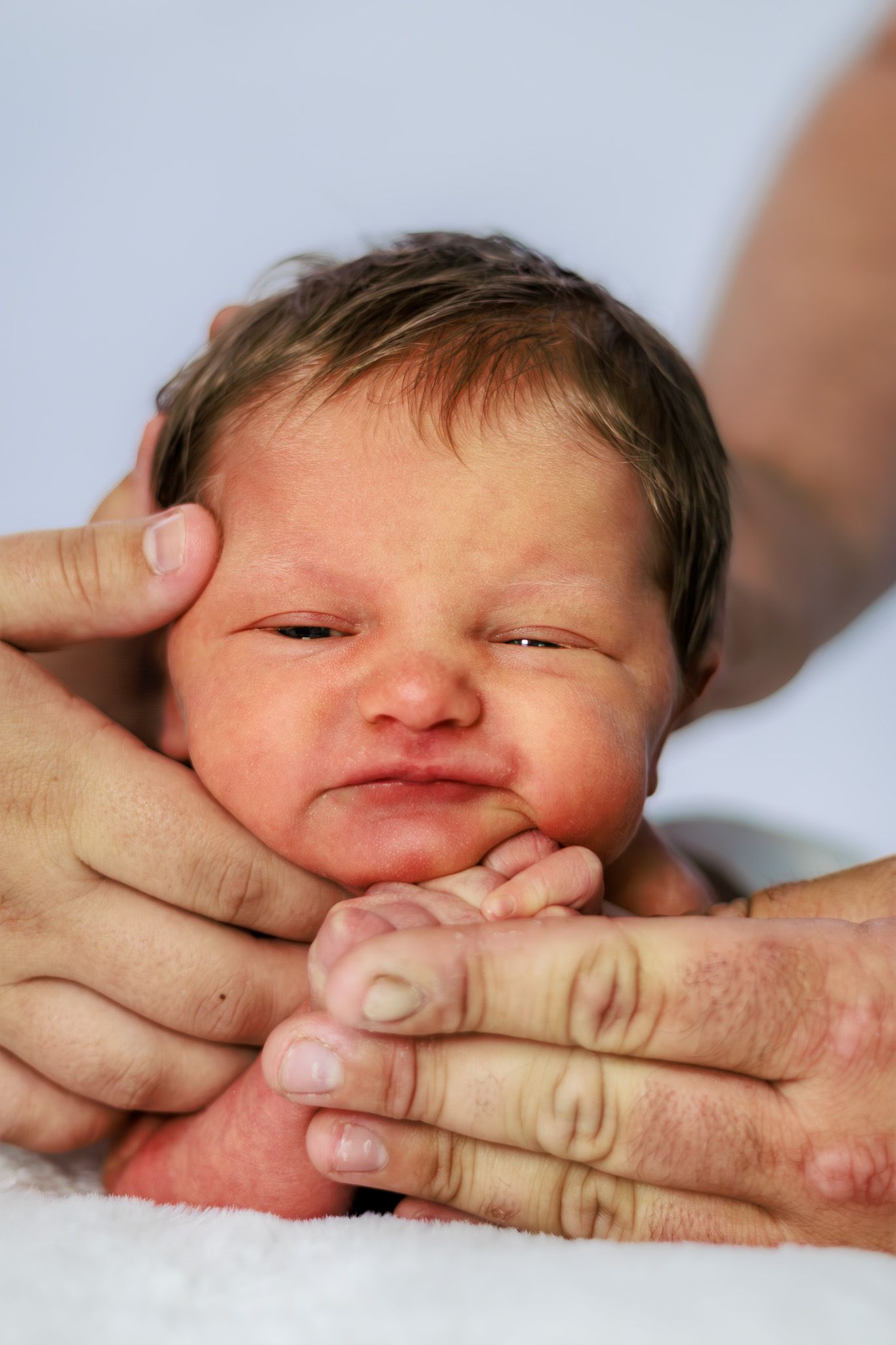 Newborn baby with dark hair resting in adult hands on a white blanket, eyes closed peacefully.