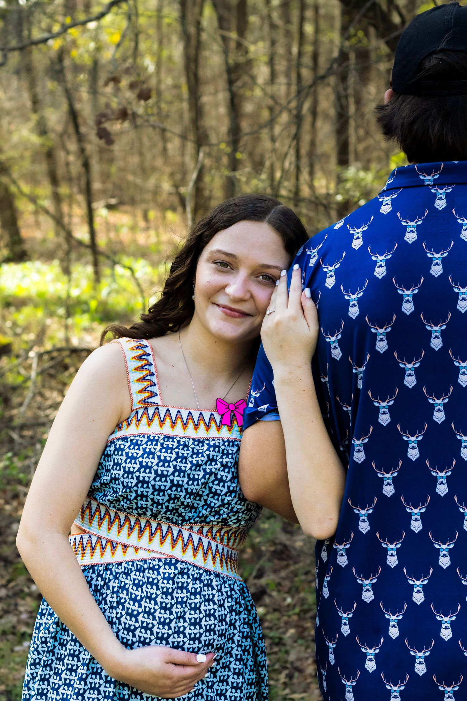 Smiling pregnant woman in patterned dress leans against man in blue deer print shirt in wooded setting.