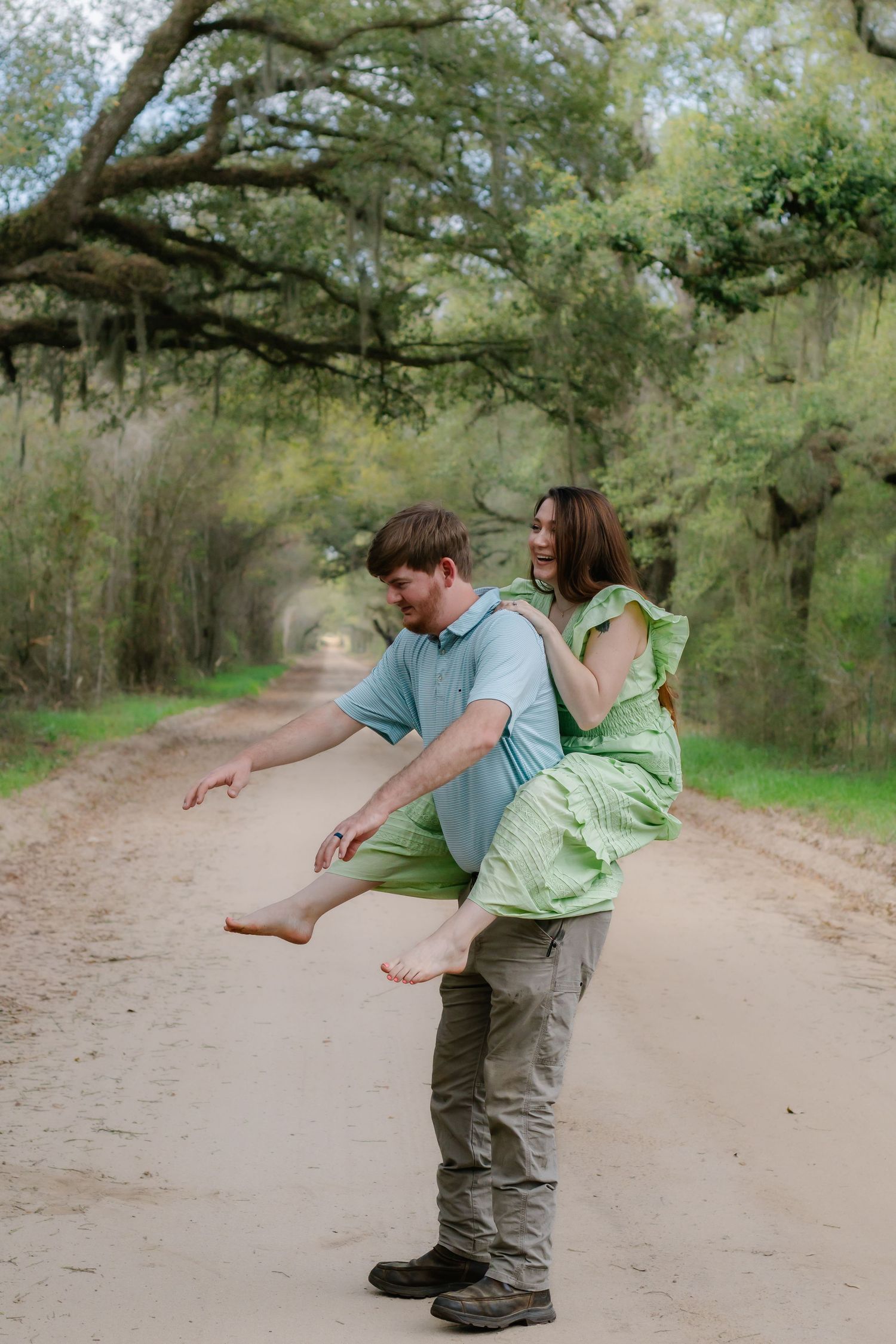 Man lifting woman in green dress on a scenic tree-canopied dirt road during engagement photos.