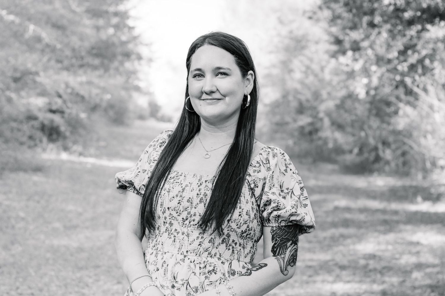 Black and white portrait of a woman with long dark hair in a floral top smiling in a park setting.