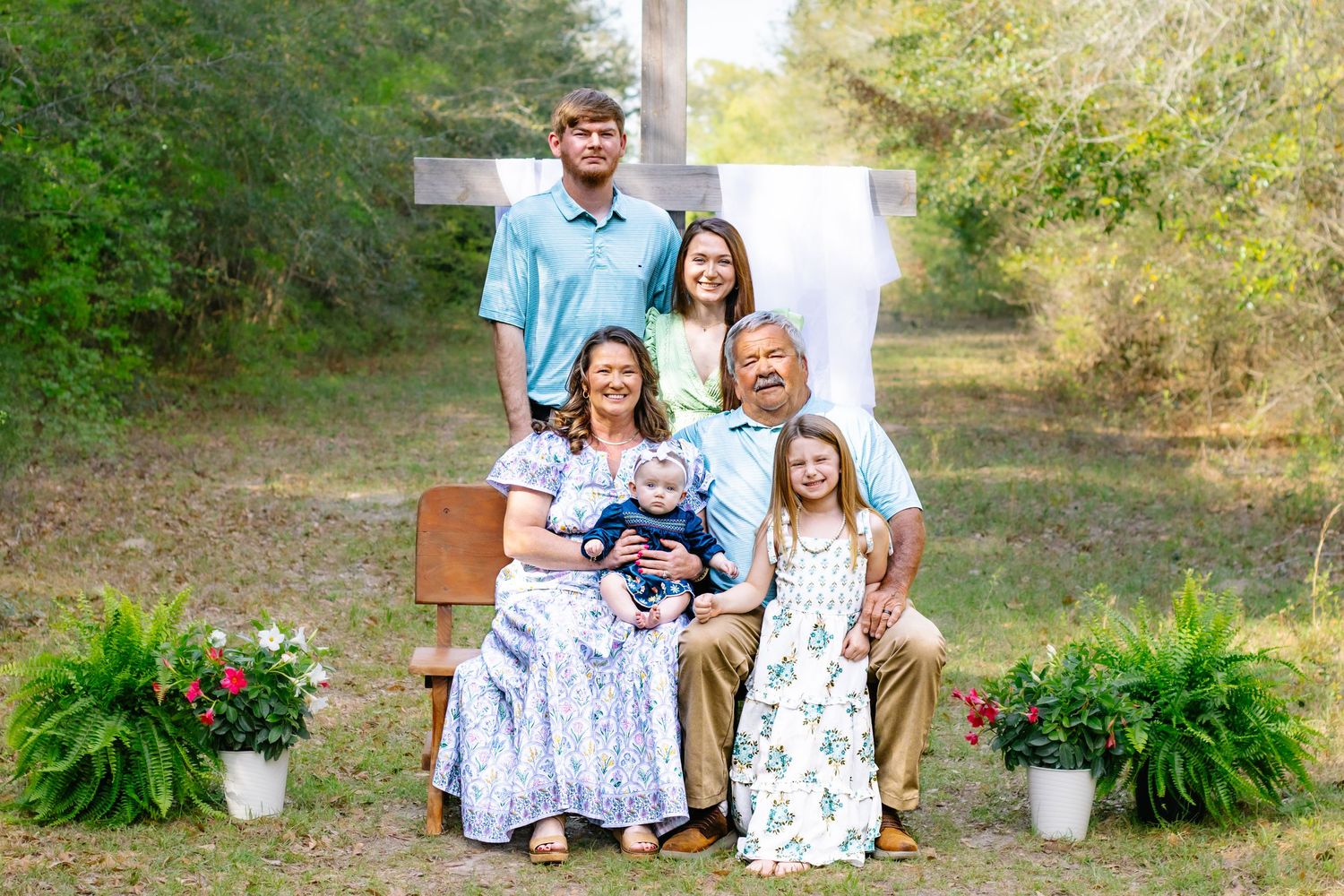 A family of six poses on a bench in front of a white cross outdoors.