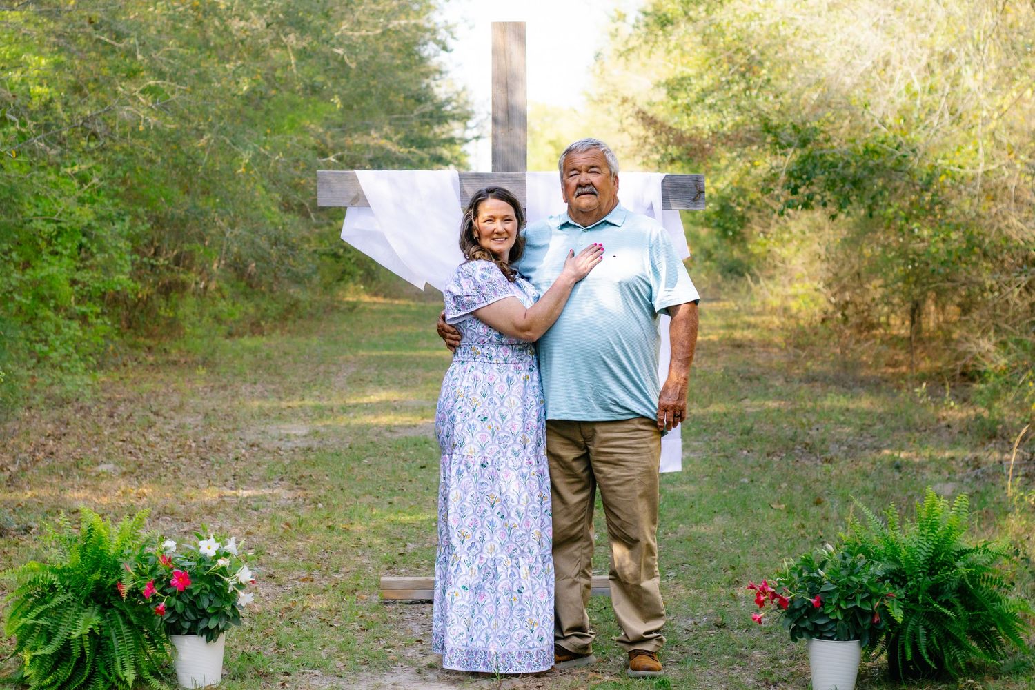 A couple embraces warmly in front of a wooden cross decorated with white fabric in a garden setting.