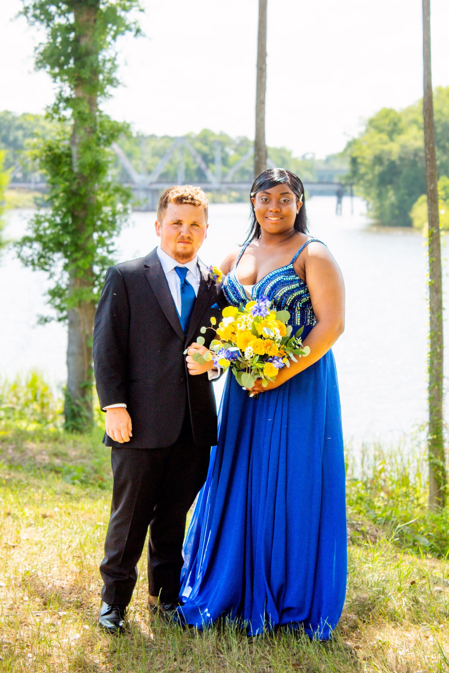 A prom couple poses outdoors near a river, the young woman in a blue gown holding a colorful bouquet.