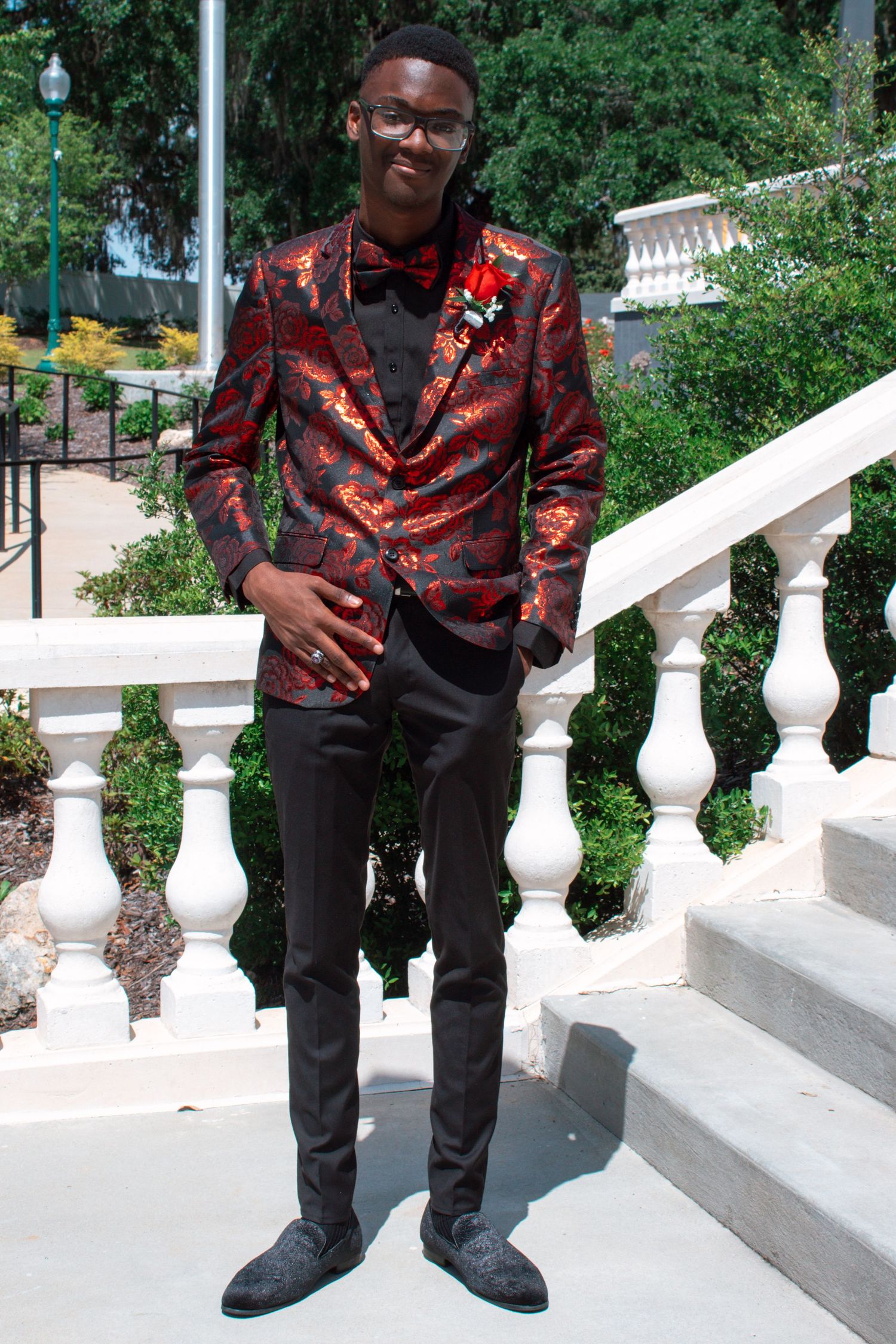 Young man in ornate red floral tuxedo jacket with bow tie poses on white steps at prom.