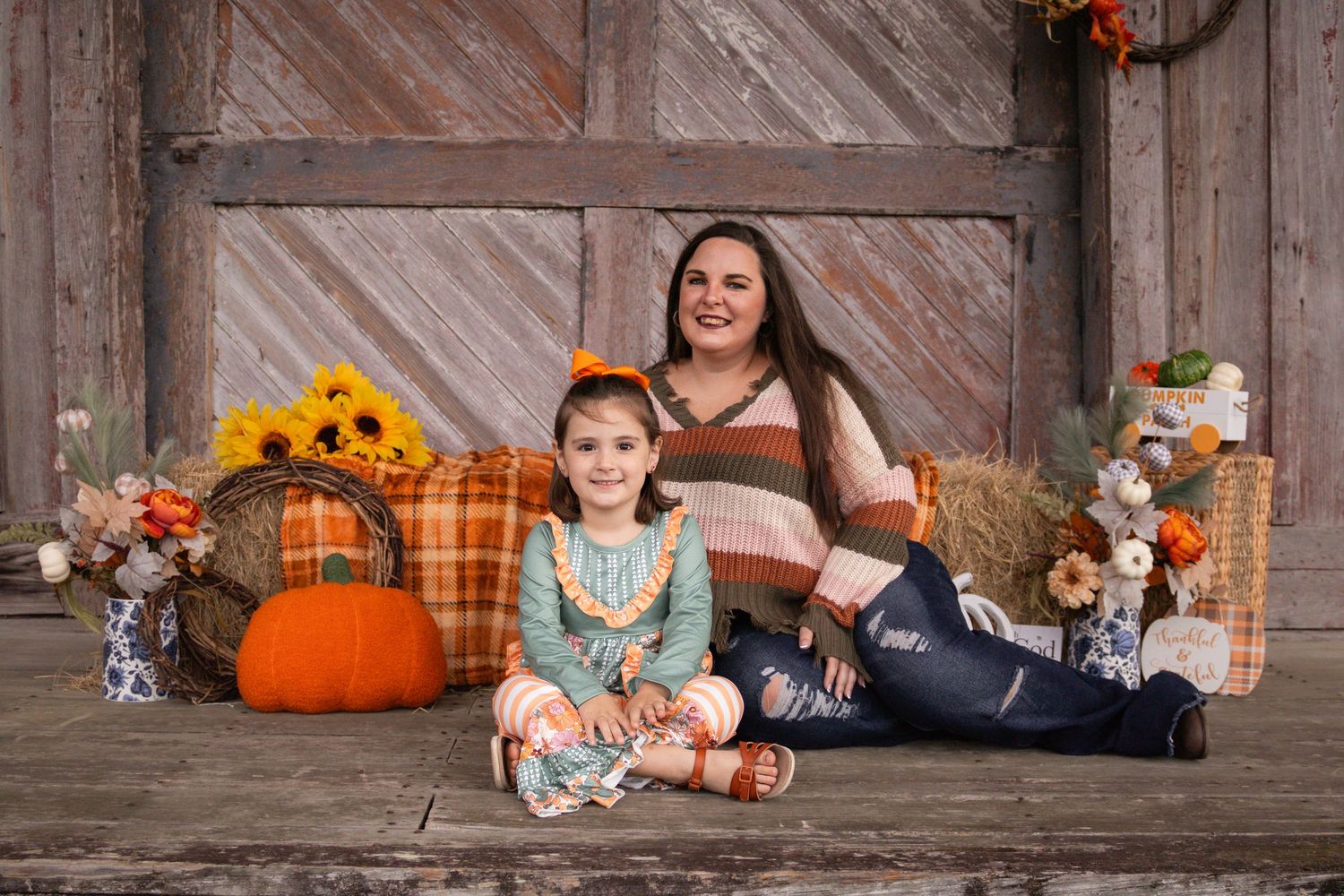Mother and daughter in fall outfits sit together surrounded by pumpkins and autumn decor by barn.