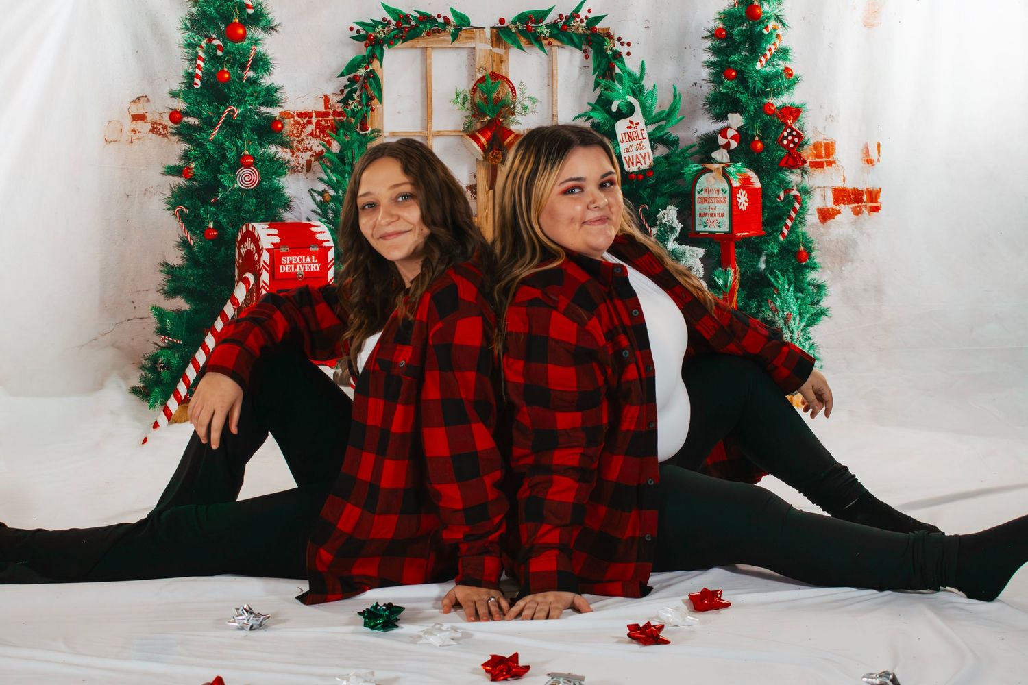 Two girls in red plaid shirts sitting back-to-back in front of Christmas tree decorations.