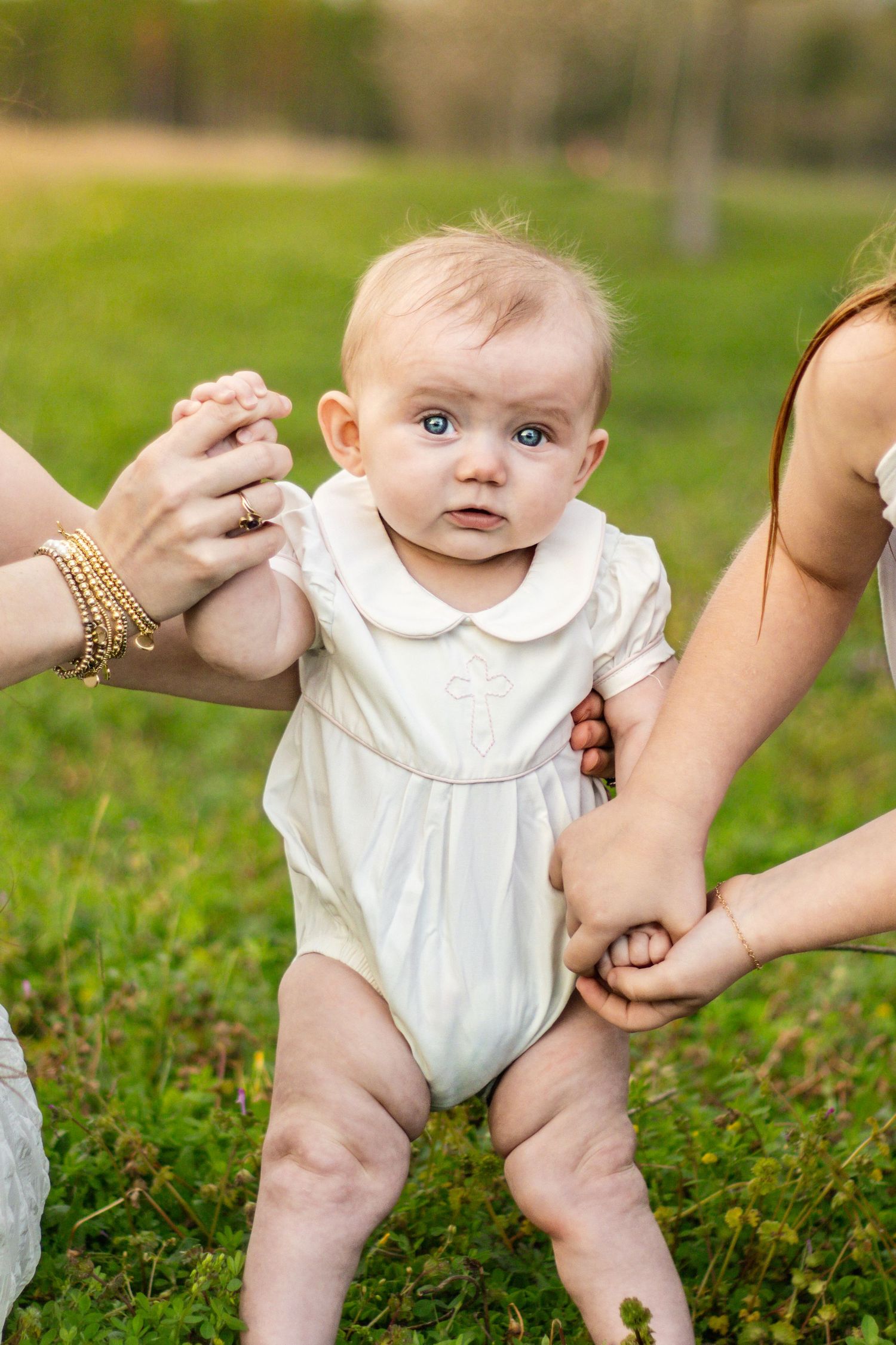 Baby in white romper supported by two adults on green grass outdoors.