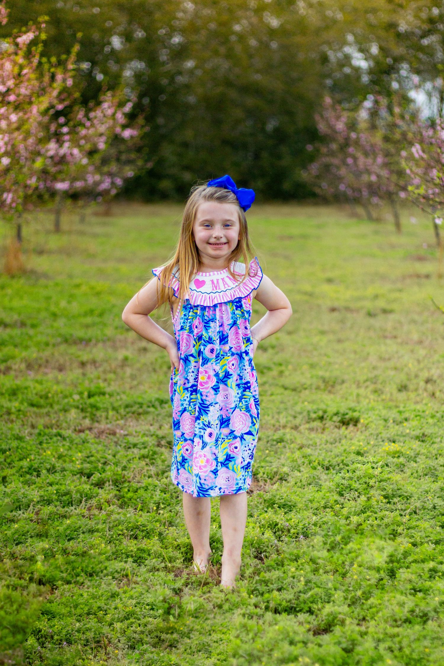 Blonde girl in blue floral dress with blue hair bow stands barefoot in green field between blooming pink trees.
