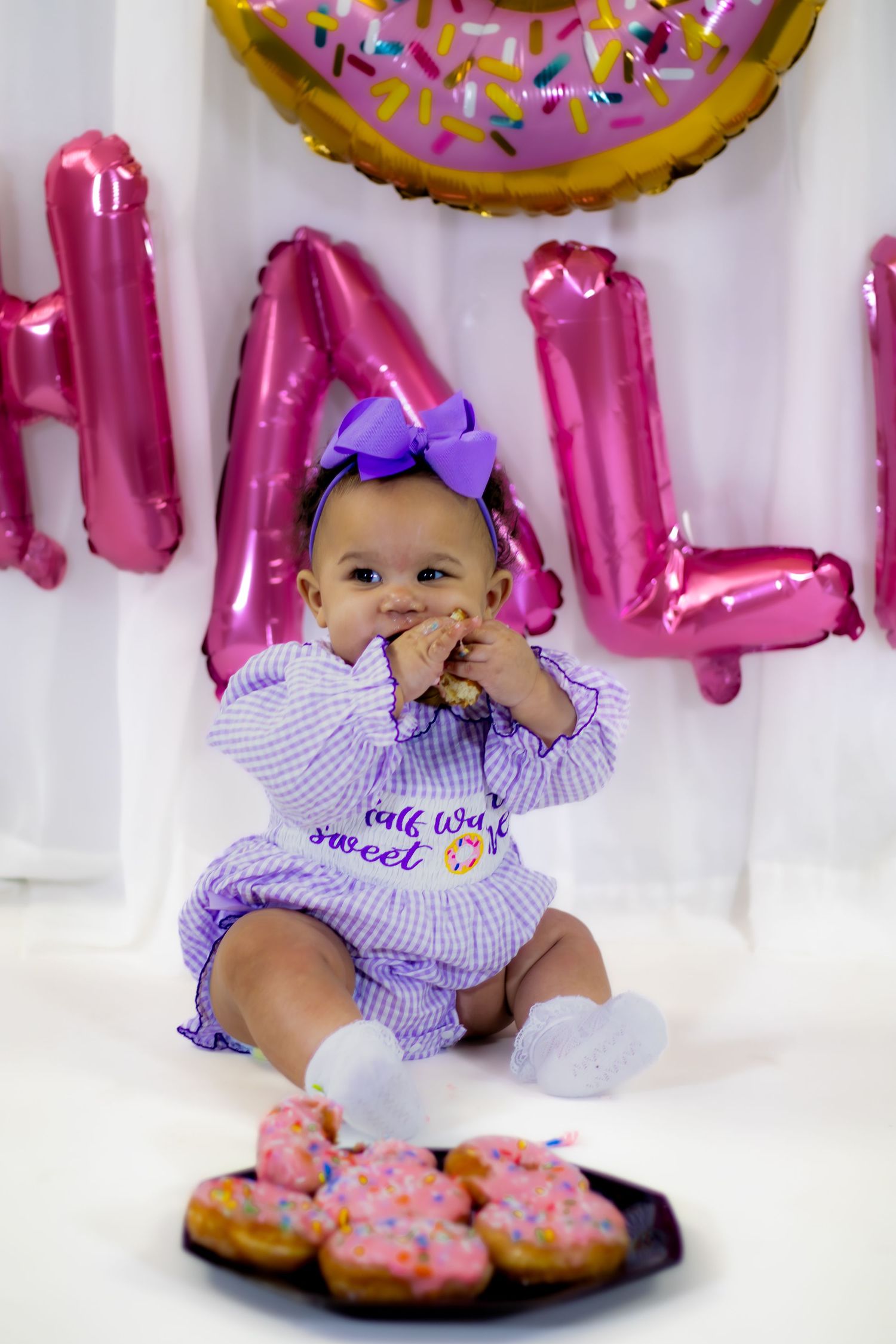 Baby girl in purple gingham romper enjoys eating donut at colorful half birthday party celebration.