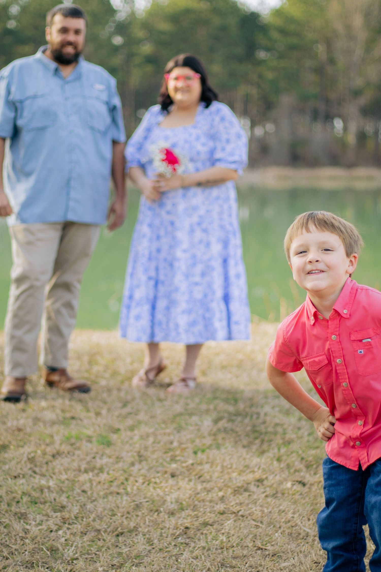 A cheerful boy in a red shirt poses confidently while his parents smile warmly behind him near a lake.