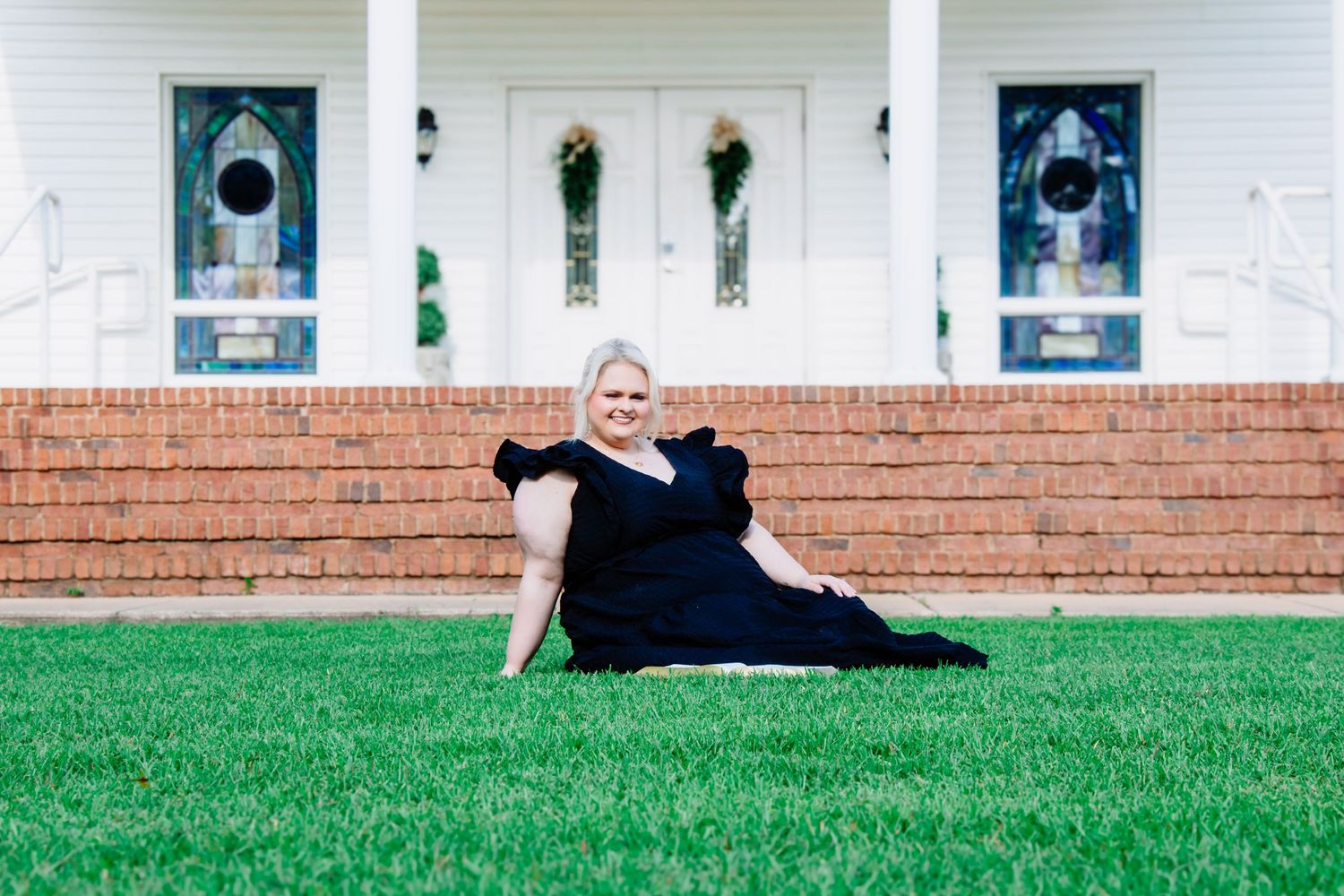 Blonde woman in black dress sitting on green grass in front of a white church with stained glass doors.