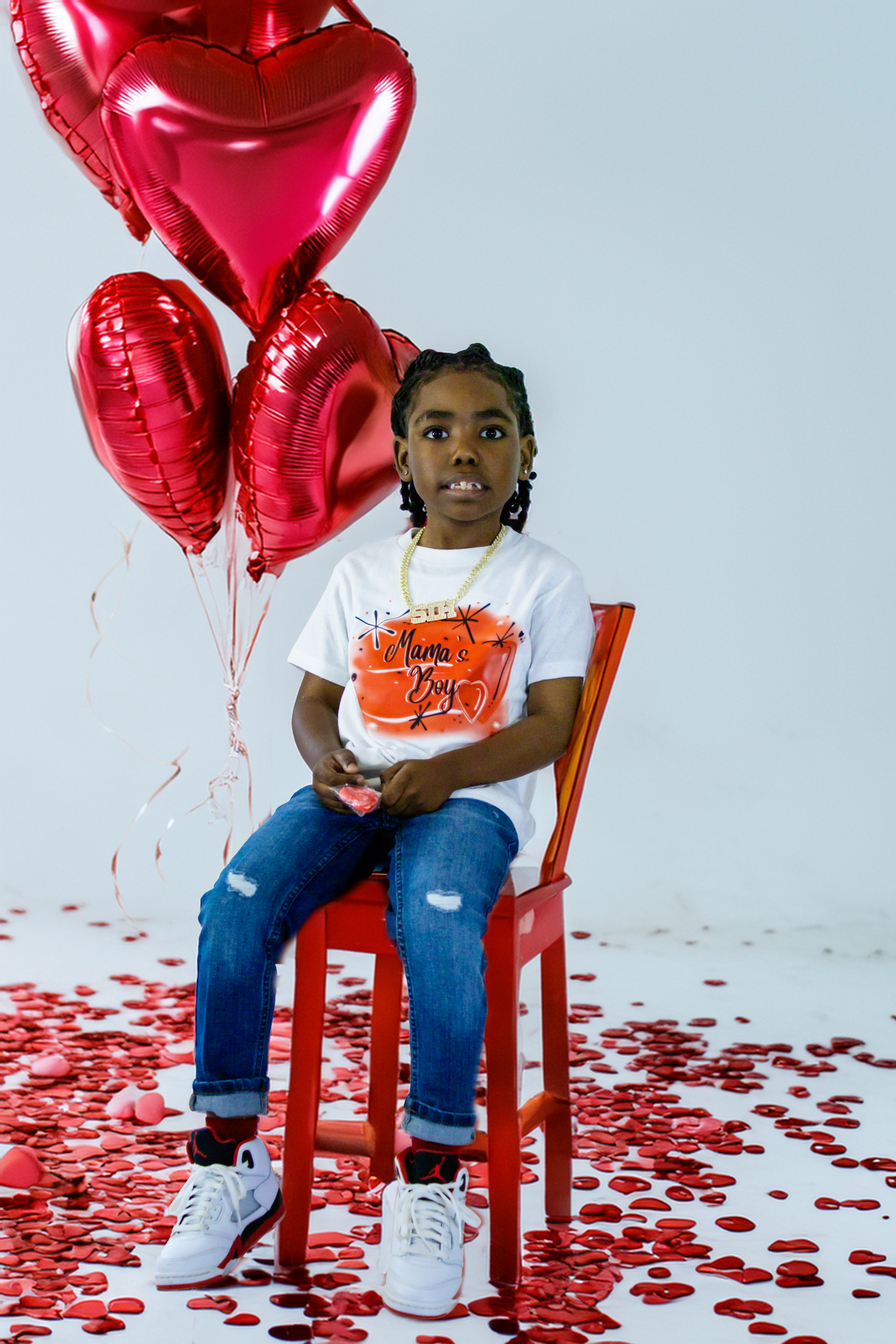 Young girl in Valentine's Day shirt sits on red chair with heart-shaped balloons and confetti.