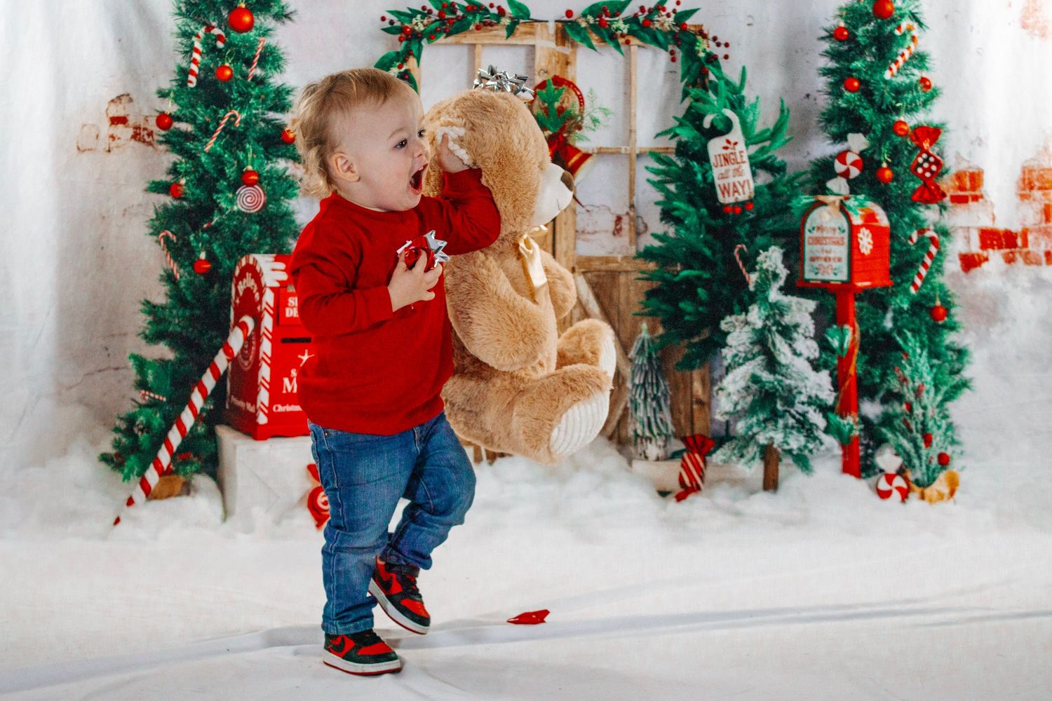 A toddler in a red sweater hugs a large teddy bear beside a decorated Christmas tree.