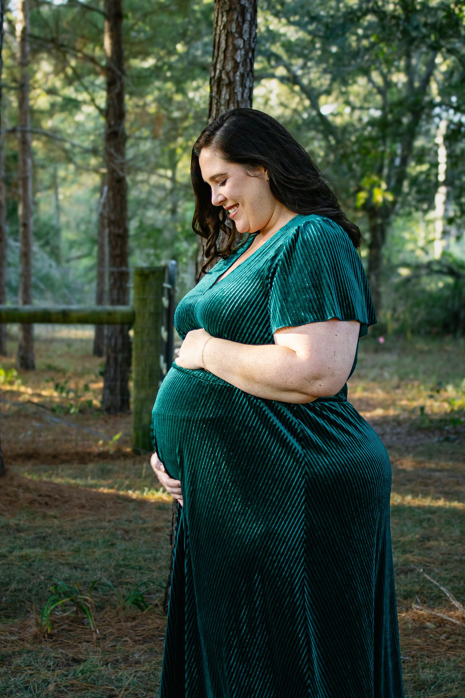 Pregnant woman in green velvet dress smiling while cradling bump in pine forest.