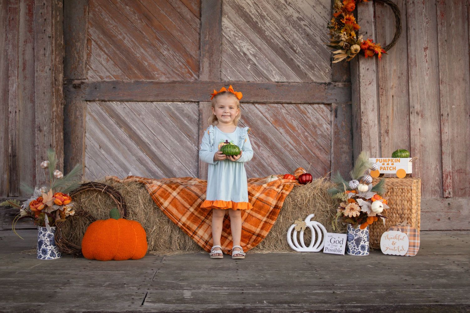 Adorable little girl in blue dress holds a pumpkin surrounded by fall harvest decorations at a barn.