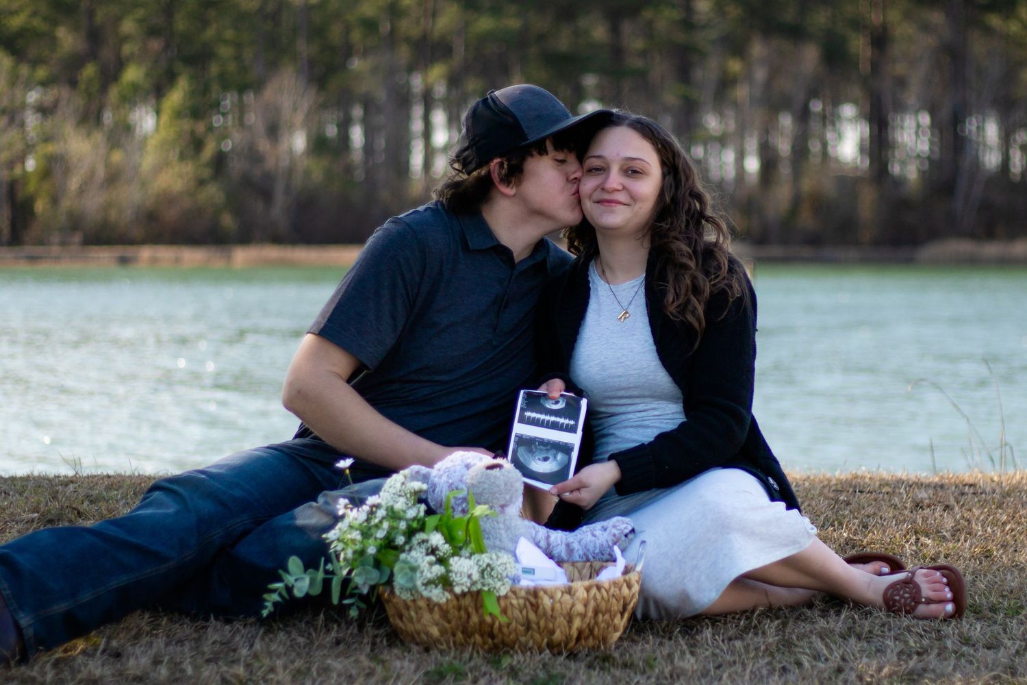 Couple sitting by lake holding ultrasound photo with flower basket.