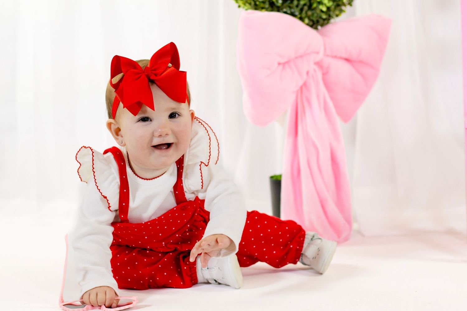 Happy baby girl in red Valentine outfit with bow headband crawls near a pink bow decoration.