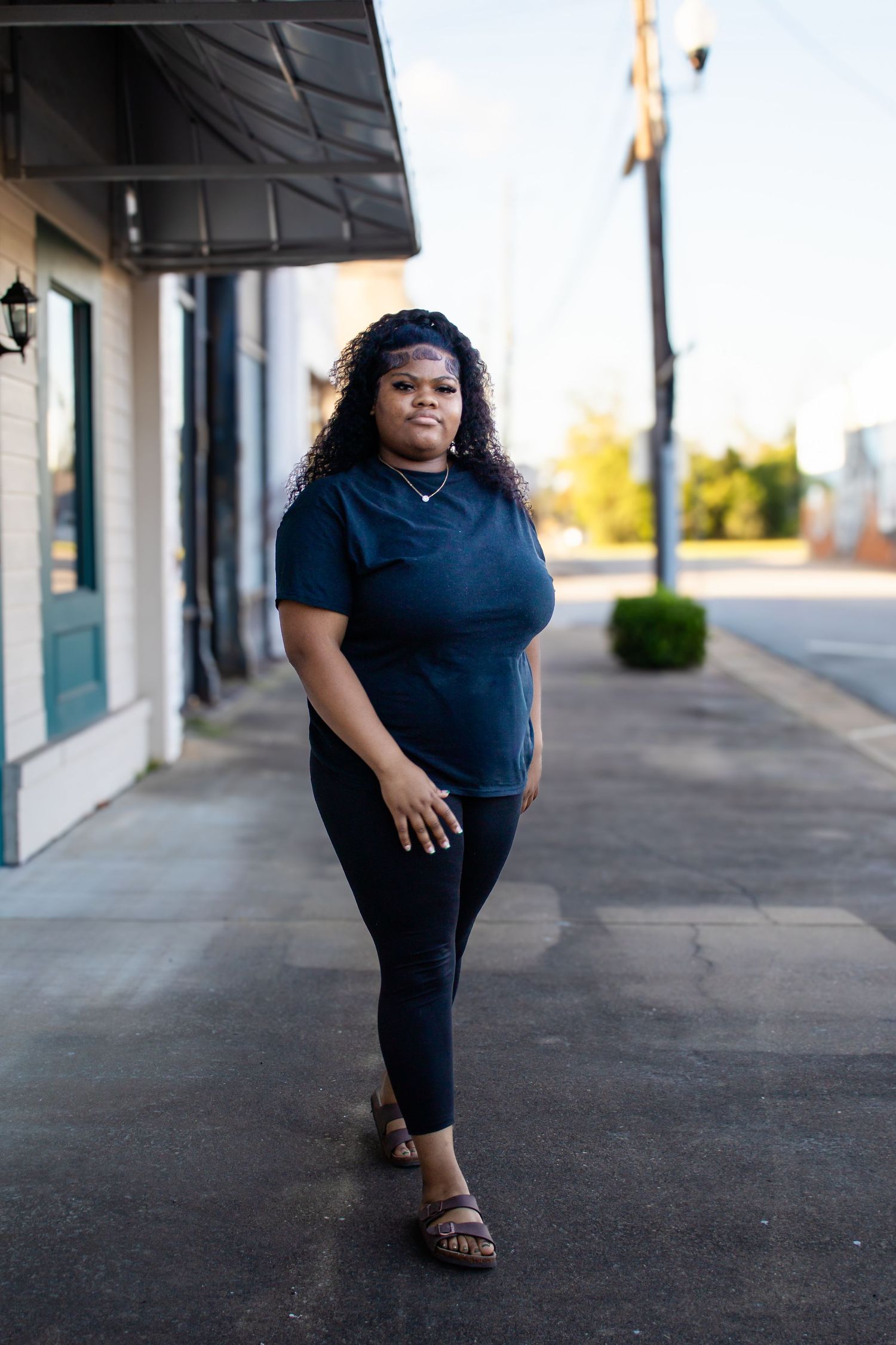Young Black woman in dark blue t-shirt and black leggings poses on a small town sidewalk.