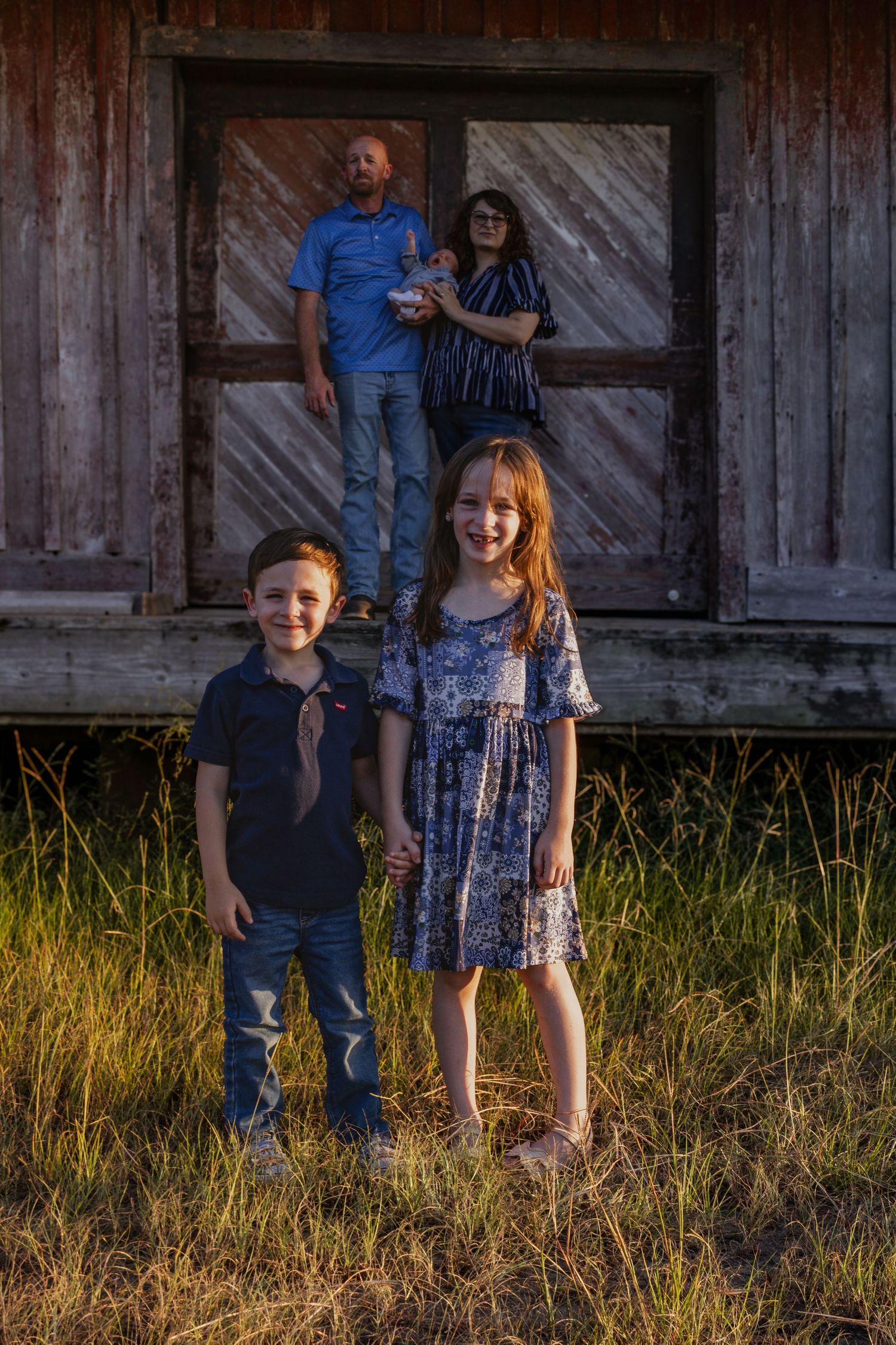 Two children holding hands in foreground while parents stand on rustic barn porch at sunset.