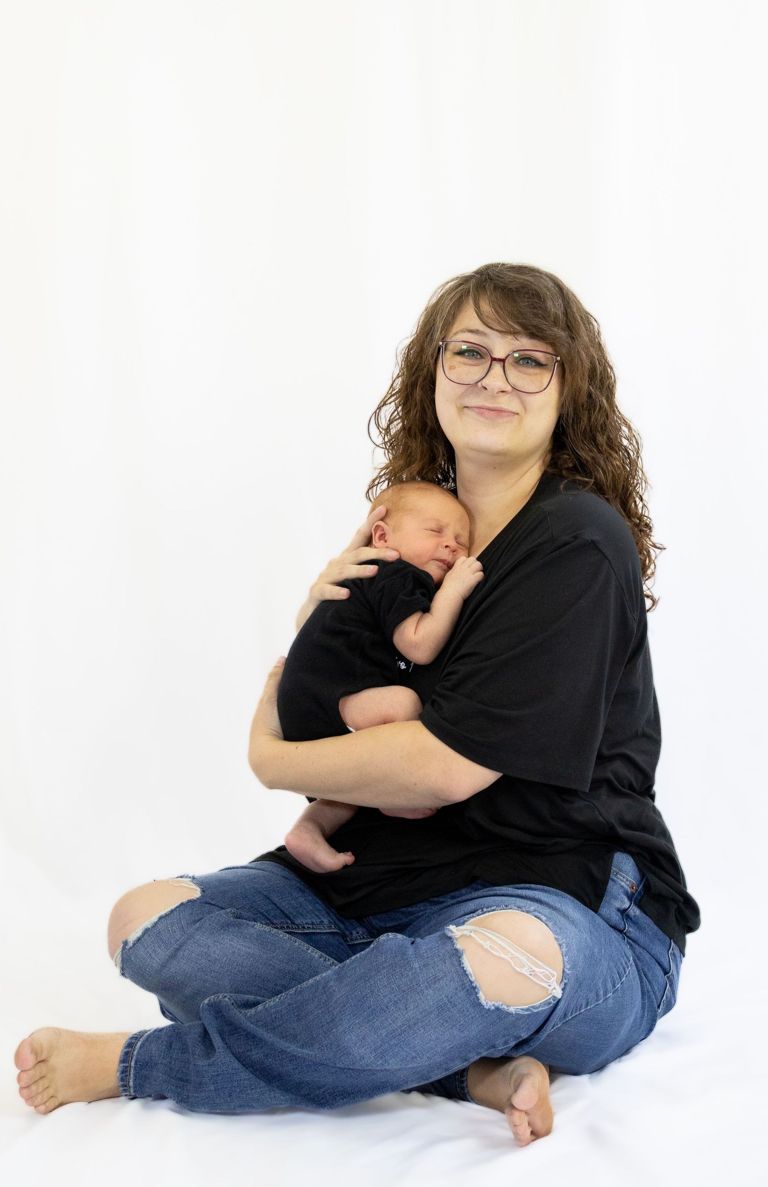 Mother with curly hair and glasses sitting cross-legged on white backdrop holding her newborn baby.