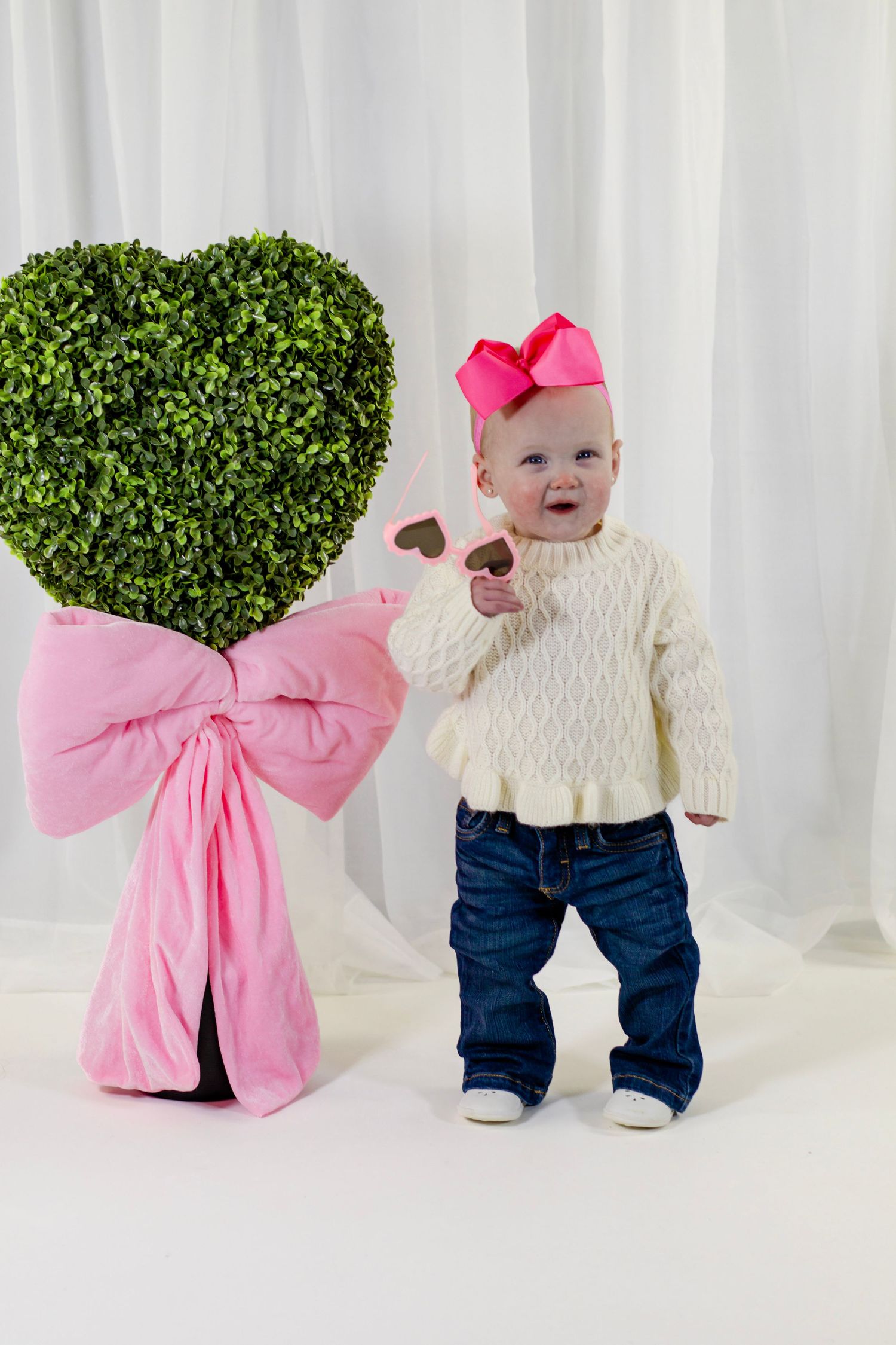 Adorable baby girl in white sweater and jeans holding heart sunglasses beside a heart-shaped topiary with pink bow.