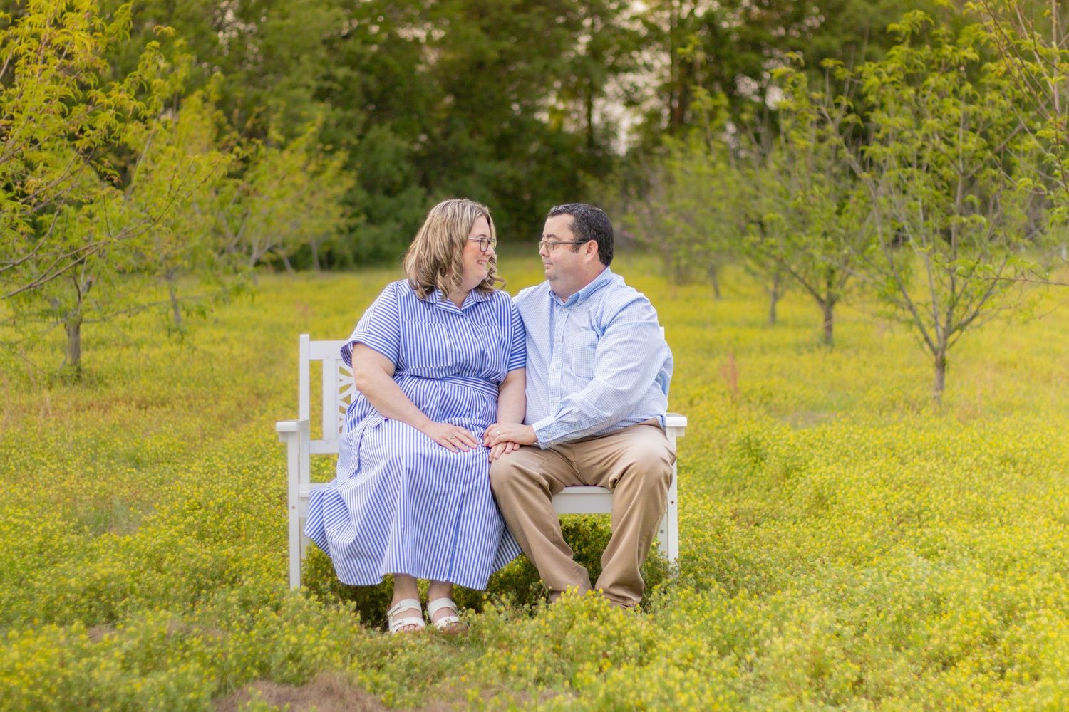 Couple sitting on white bench in yellow wildflower field gazing lovingly at each other.