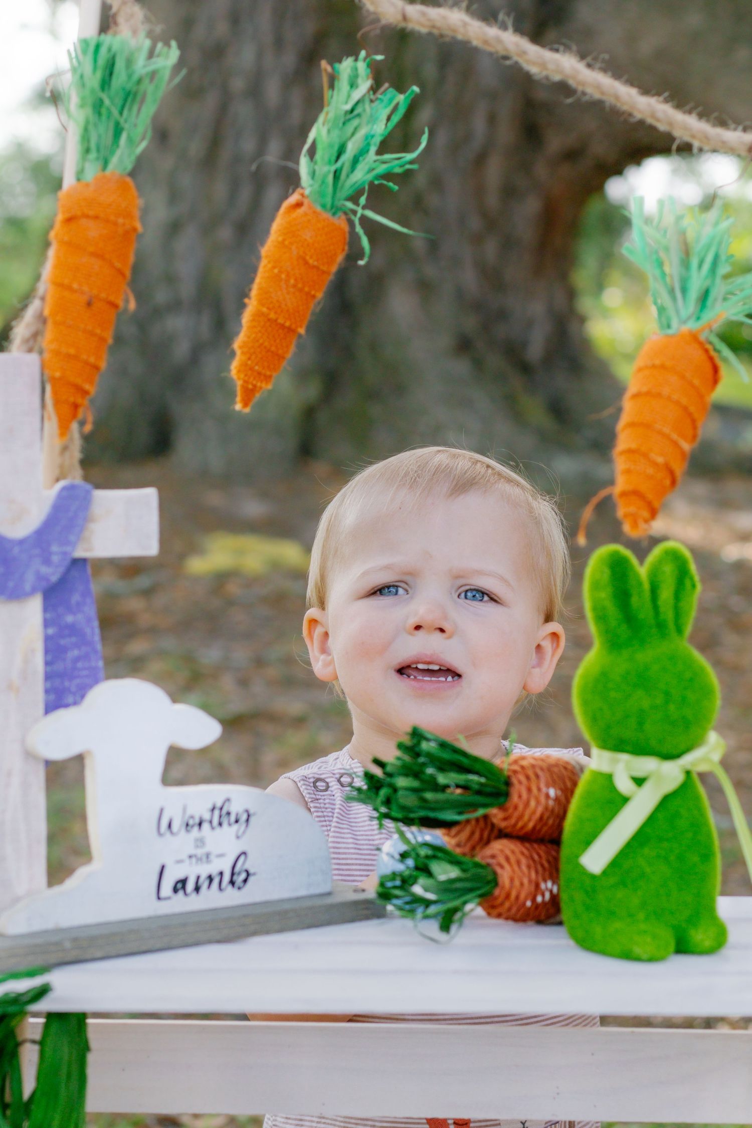 Blond toddler boy peeking over white table with green Easter bunny and hanging carrot decorations outdoors.
