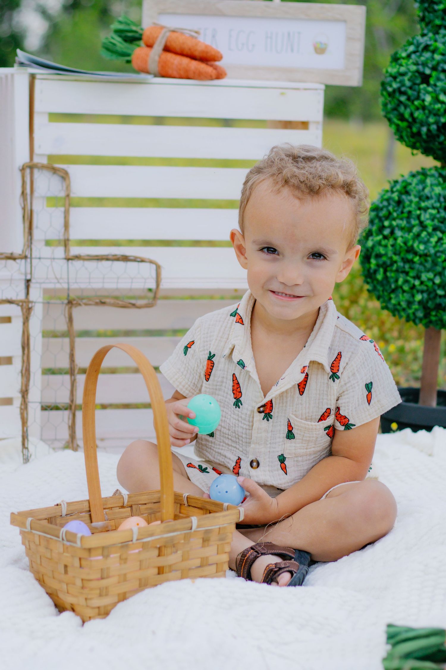 Smiling toddler boy in carrot-print romper sits cross-legged holding Easter egg beside a wicker basket outdoors.