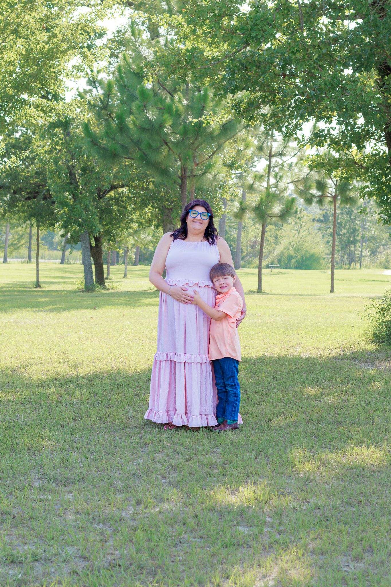 Pregnant woman in pink maxi dress stands with young boy in park setting surrounded by lush green trees.