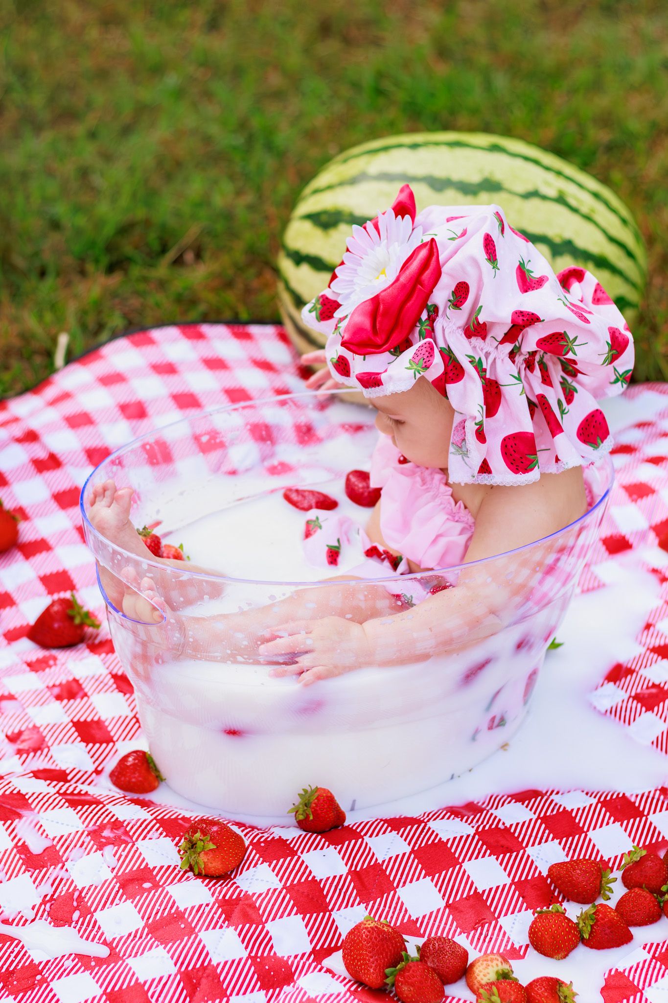 Baby girl in strawberry dress plays in milk bath with strawberries on red checkered blanket outdoors.