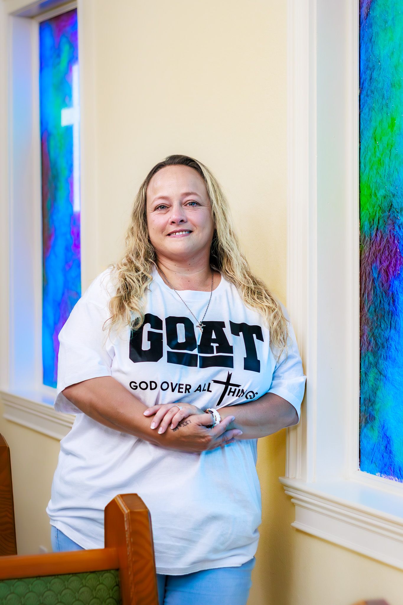 Woman in GOAT God Over All Things shirt leaning against wall near colorful stained glass windows in church.
