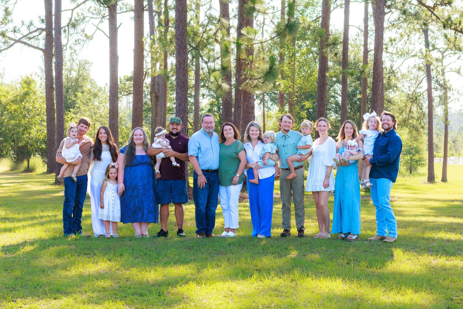 Large extended family group portrait outdoors among tall pine trees on green grass in natural light.