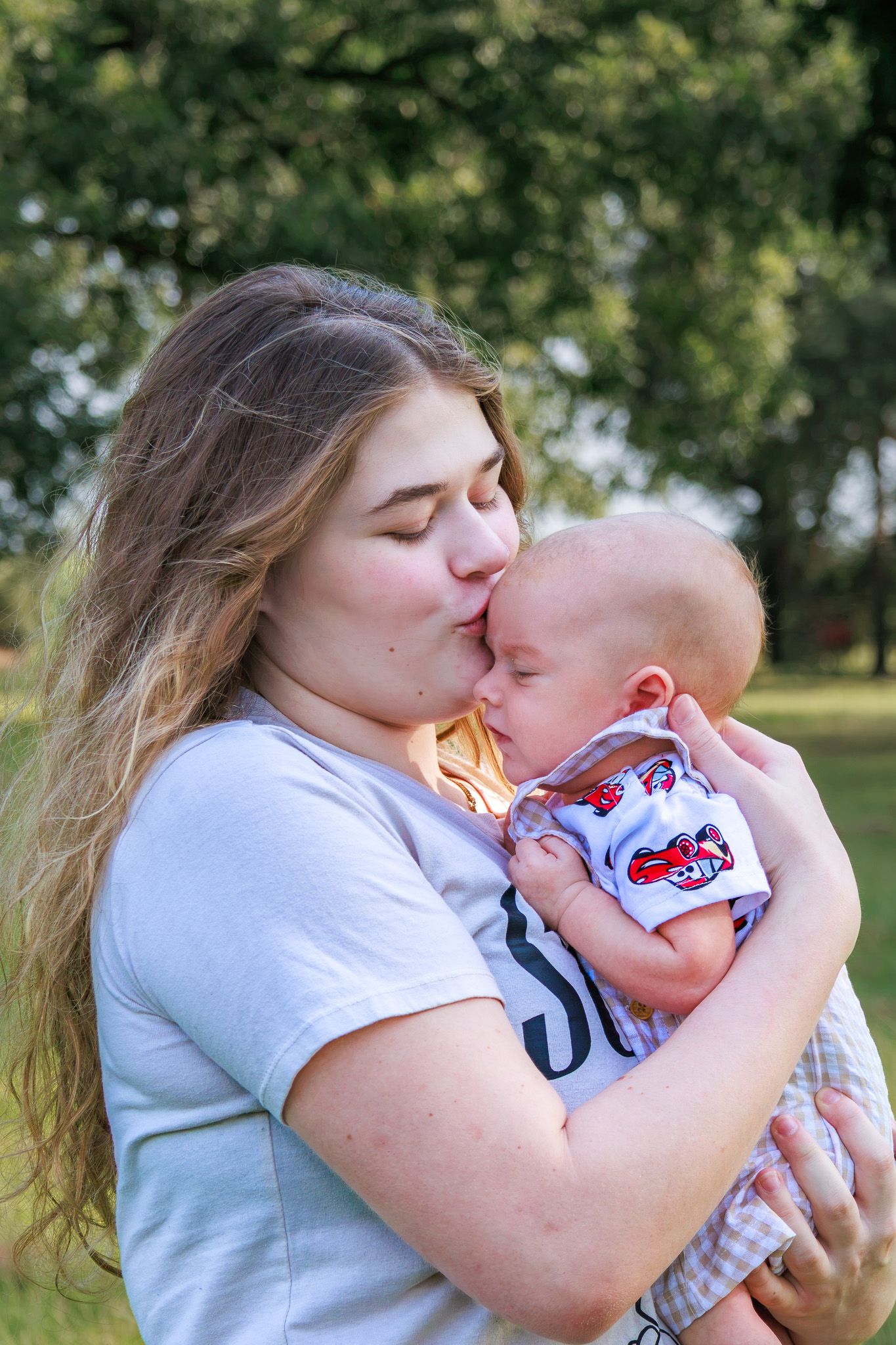 Young mother kissing her newborn baby outdoors in a green park setting on a warm sunny day.