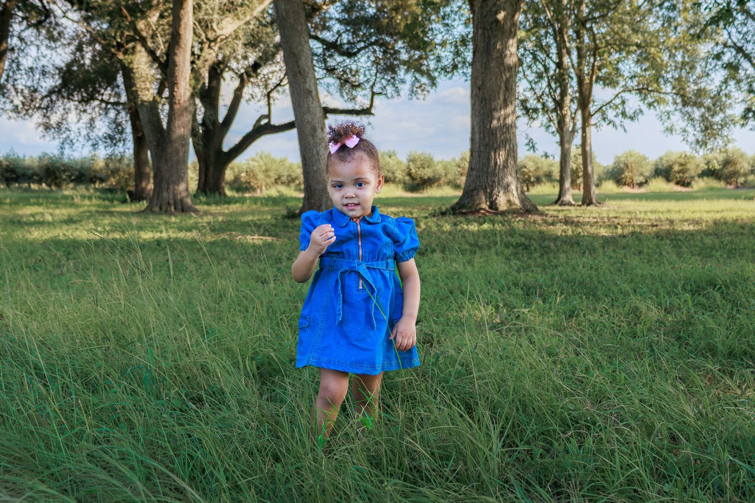 Young girl in blue dress running through green grass field with large trees in background.