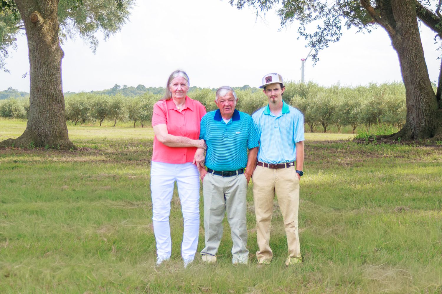 Three people smiling outdoors on a green field with trees, wearing casual polo shirts on a sunny day.
