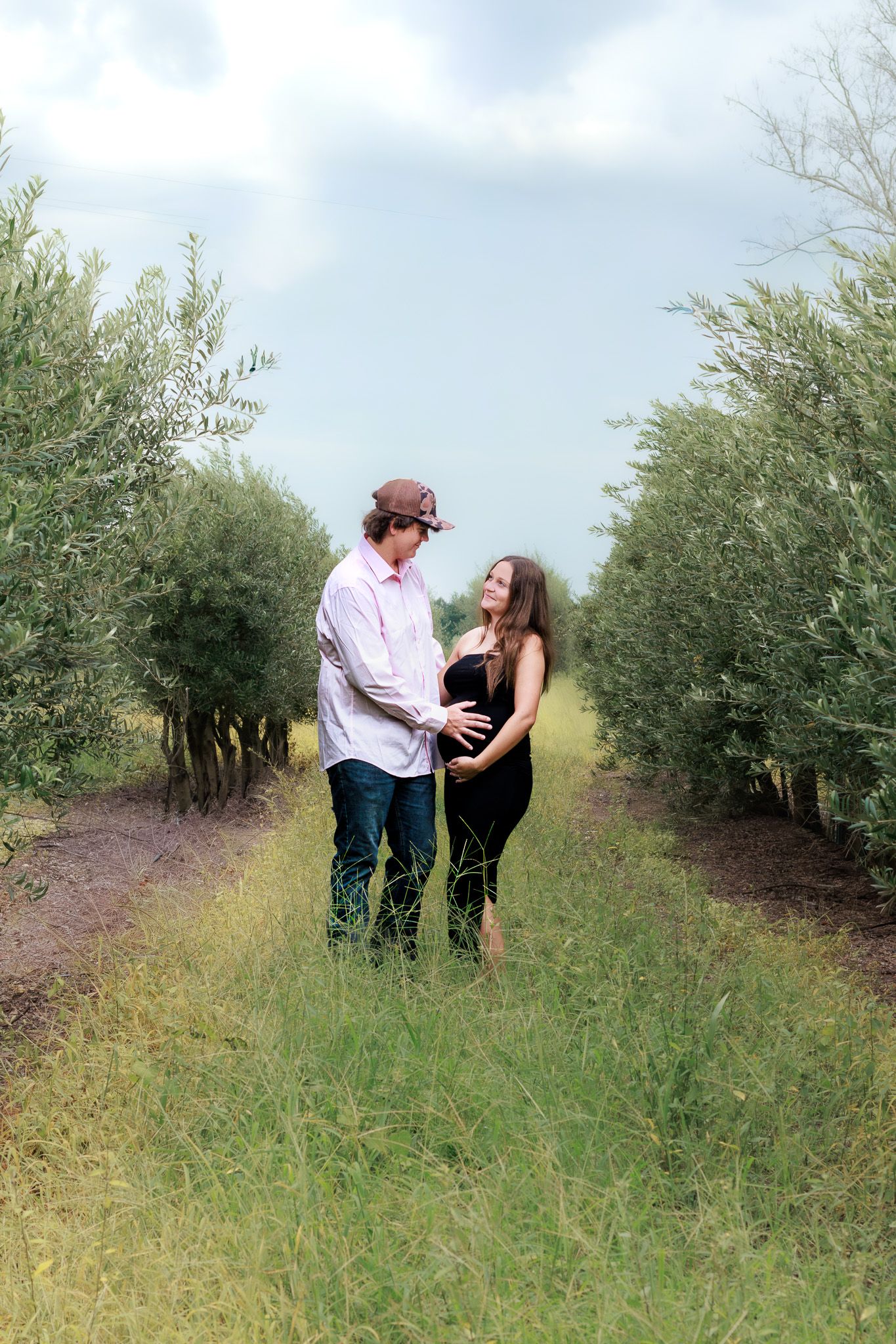Couple sharing a tender moment in a lush green orchard pathway, woman in black dress with visible baby bump.