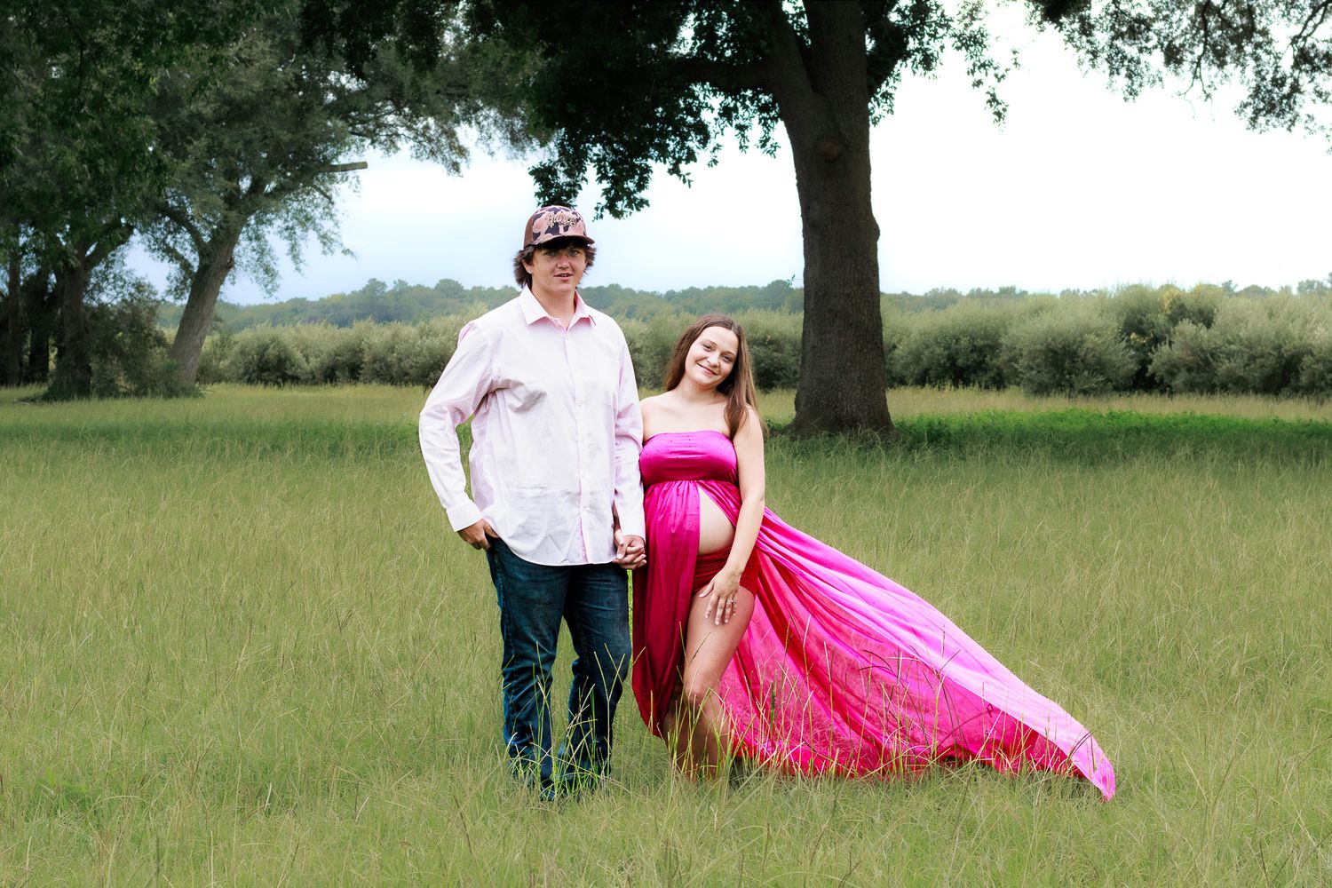 Pregnant couple posing in a grassy field, woman in elegant pink maternity gown with flowing train.
