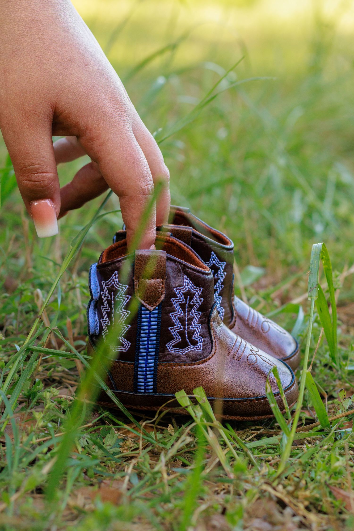 A hand with manicured nails picks up a tiny brown cowboy boot with blue embroidery from green grass.
