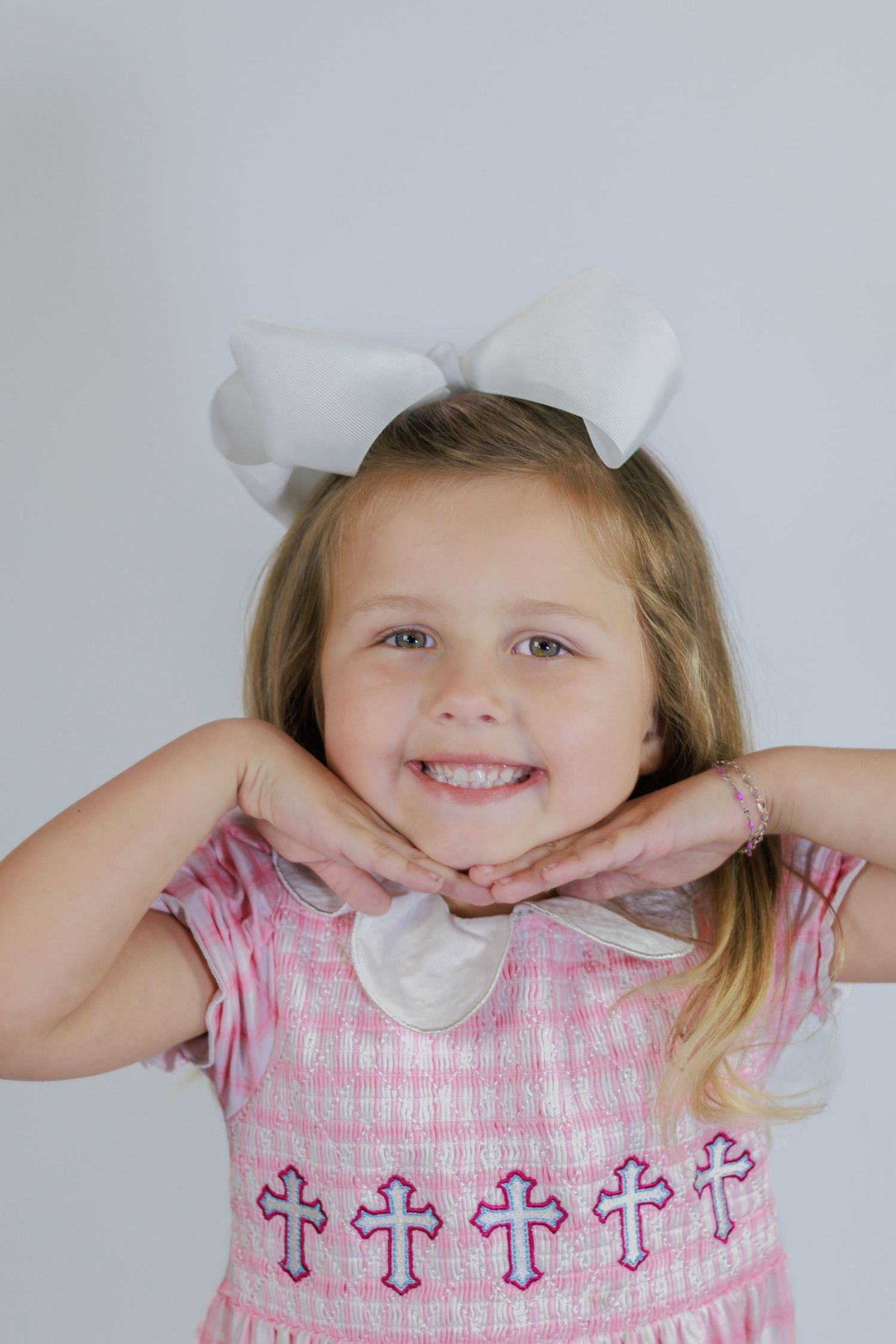 Smiling young girl in pink cross-embroidered dress with large white hair bow posing with hands under chin.