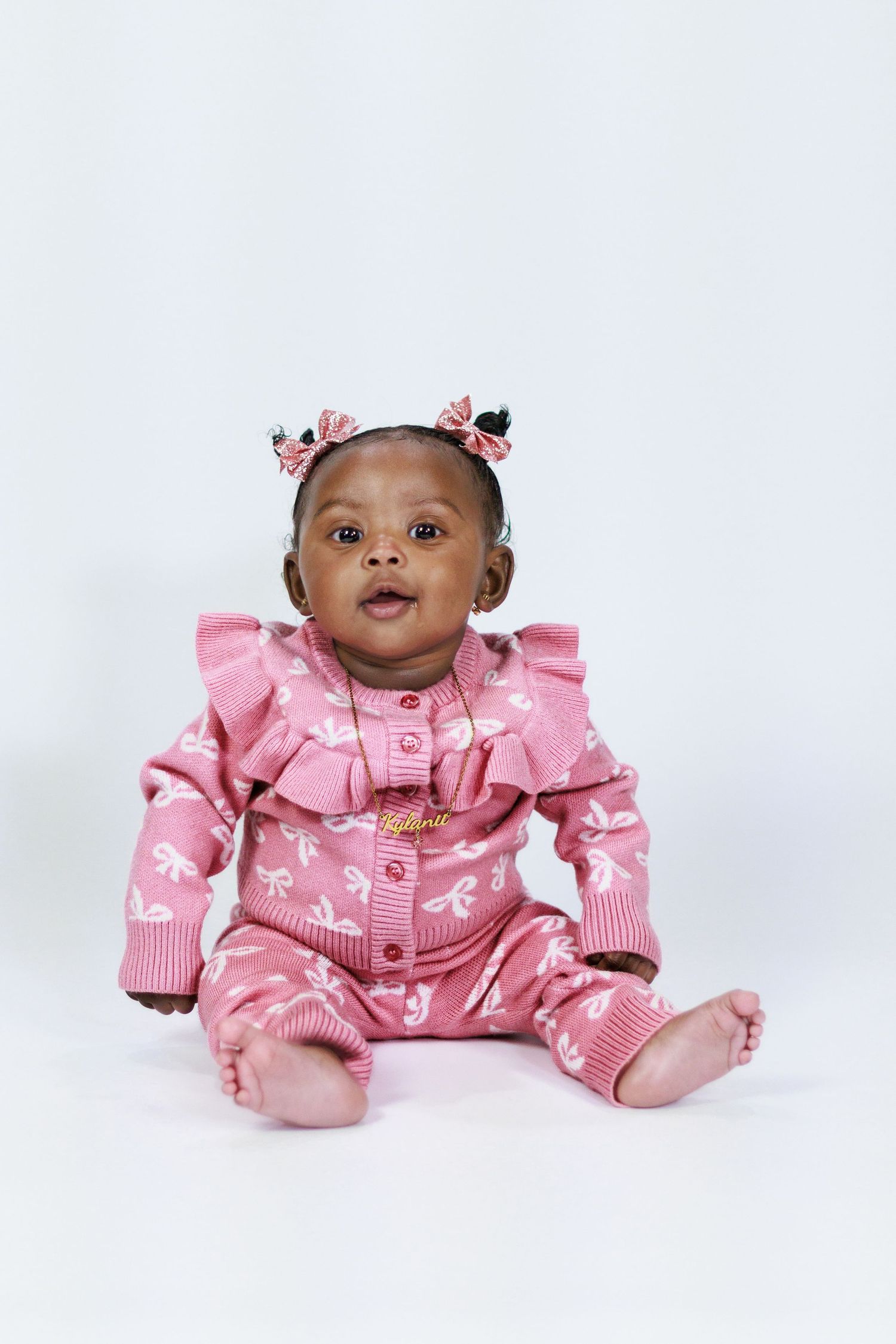 Cute baby girl in pink ruffled knit outfit with bow pigtails sitting on white background.