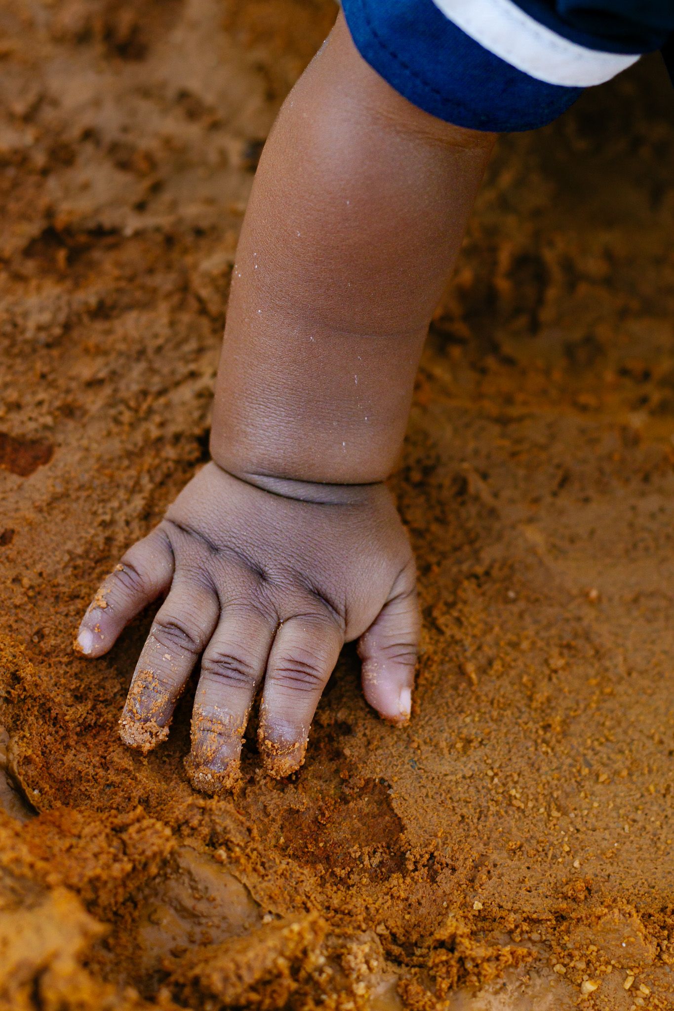 Close-up of a small child's hand pressing into reddish-brown sandy dirt outdoors.